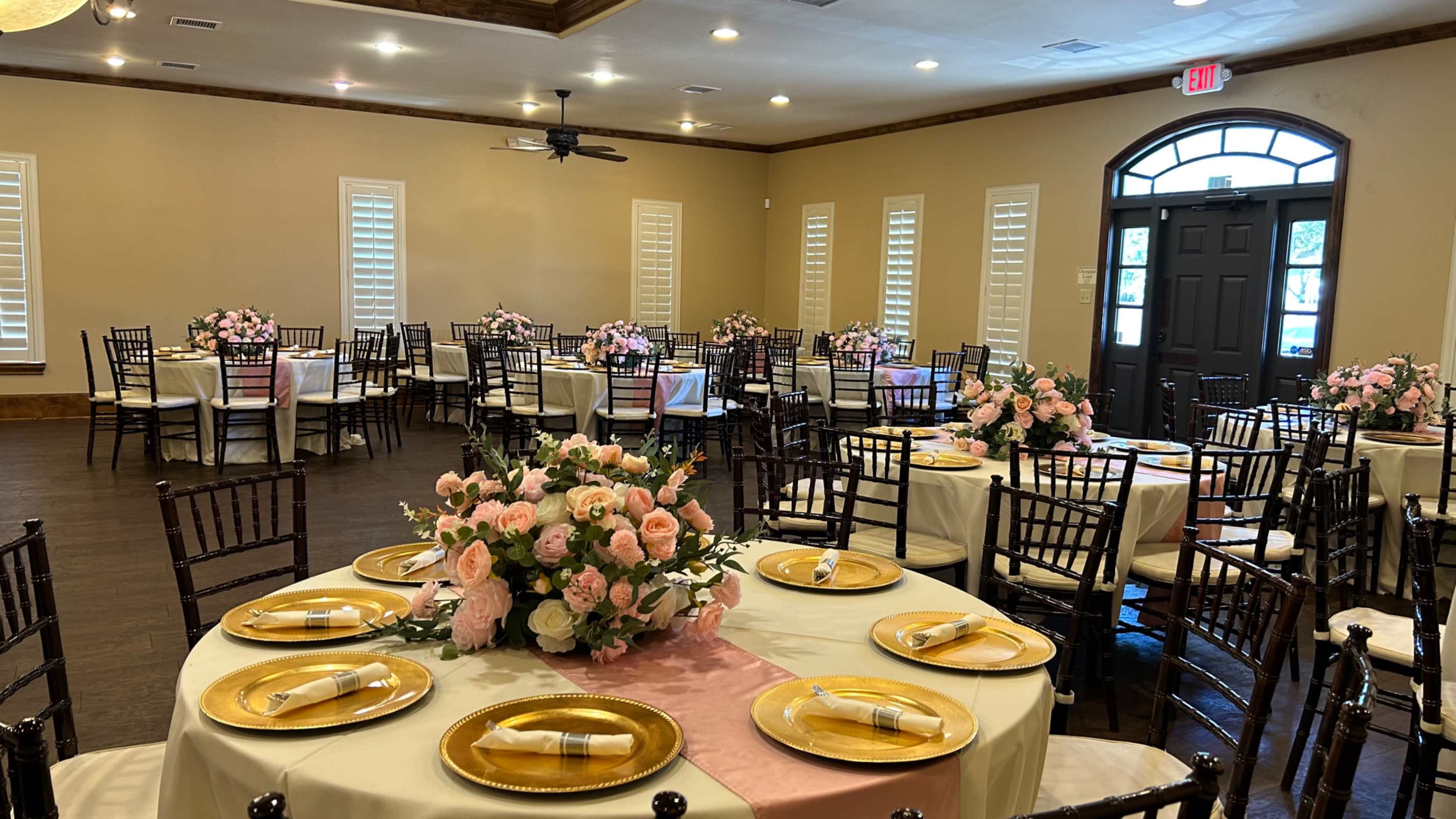 A banquet hall arranged for an event, featuring round tables with floral centerpieces, gold-rimmed plates, and chairs arranged around them.