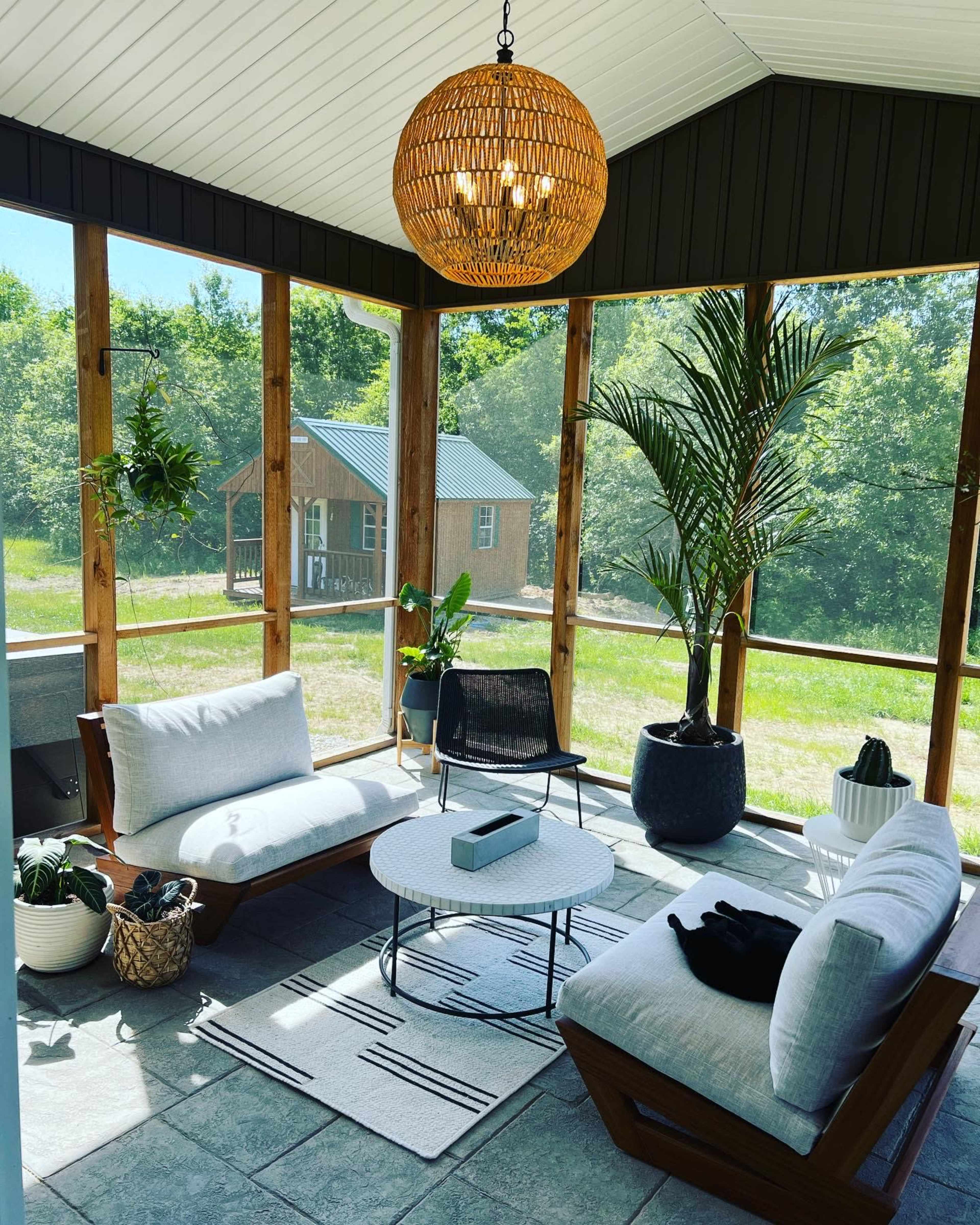 The image shows a sunlit screened-in porch decorated with a large pendant light, comfortable seating, and various houseplants.