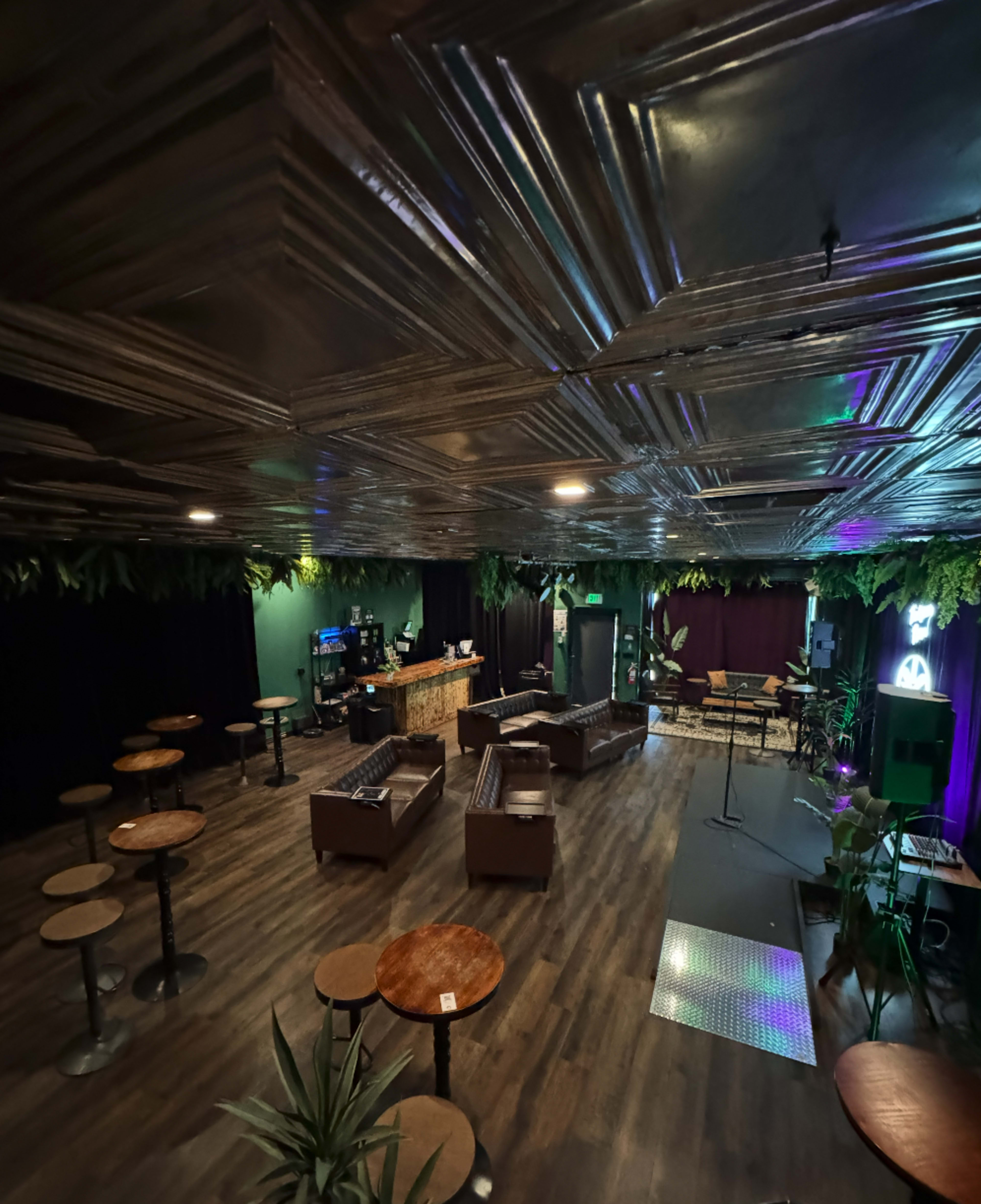 The image shows a dimly lit lounge area with wooden flooring, arranged seating, and a small stage, decorated with plants along the ceiling.