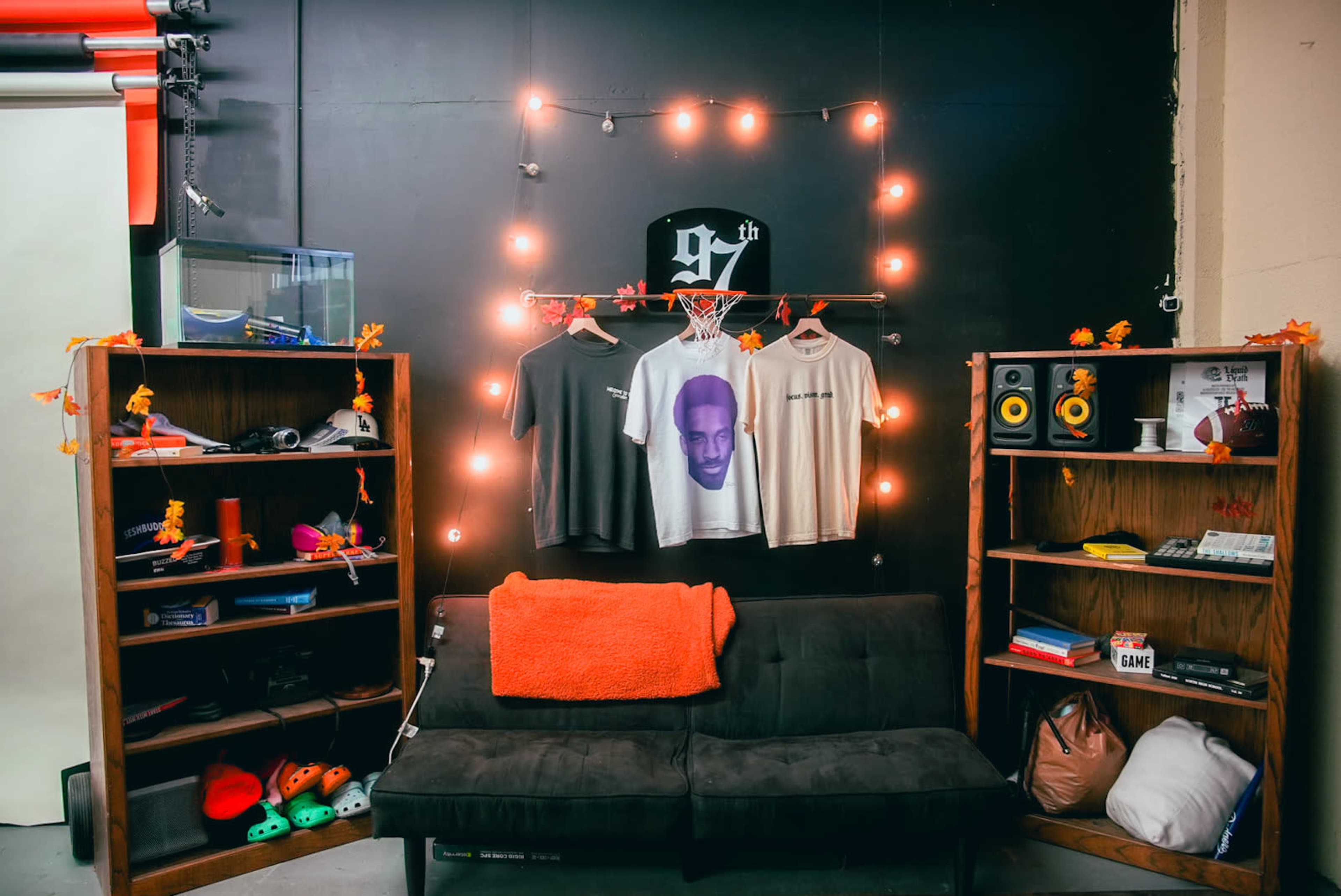 A cozy room with a black wall adorned with string lights, featuring two wooden shelves filled with various items, a black couch, and three hanging T-shirts.