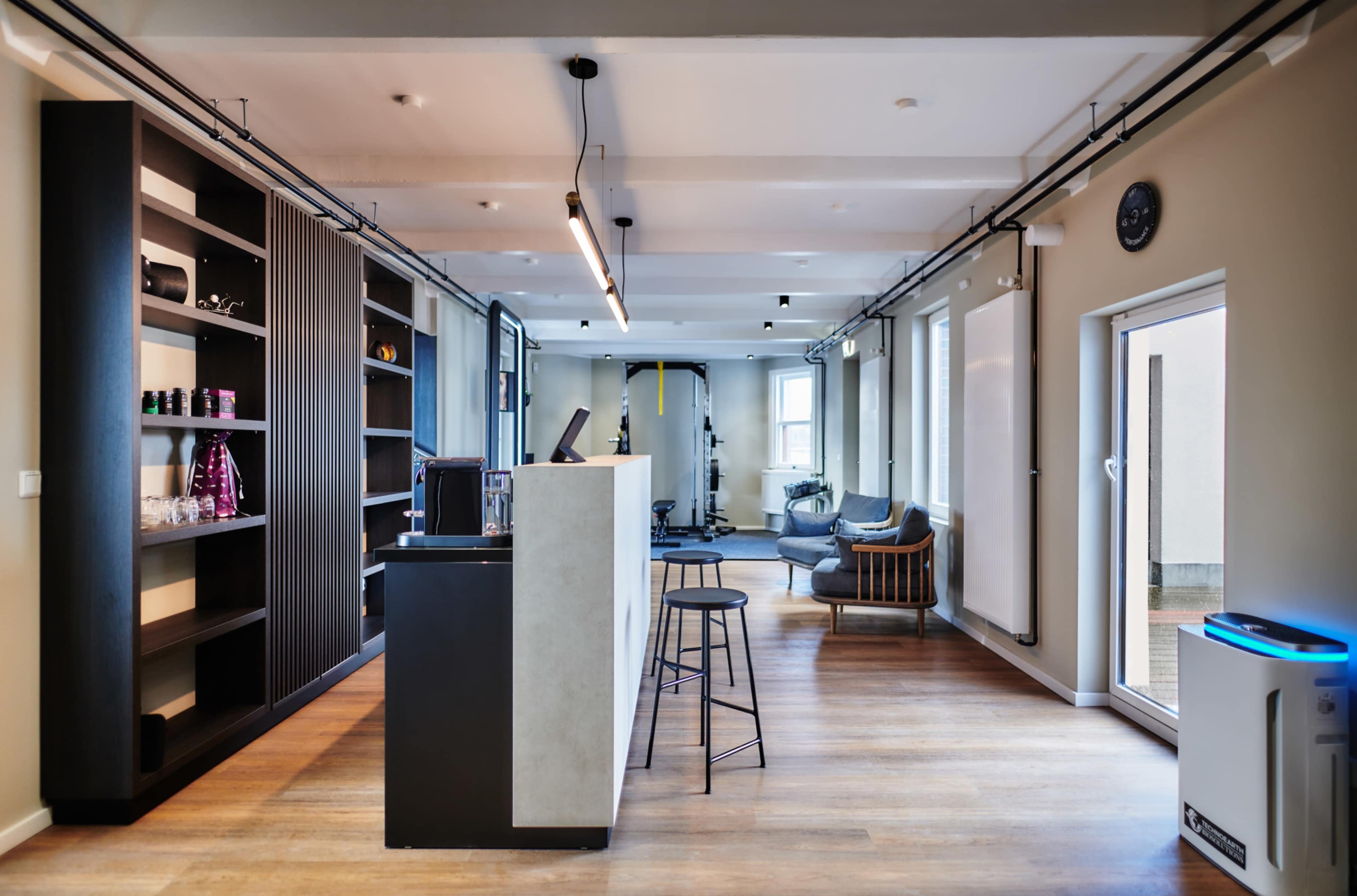 The image shows a modern, open office space with a reception area, bookshelves, and a seating area, featuring large windows and wooden flooring.