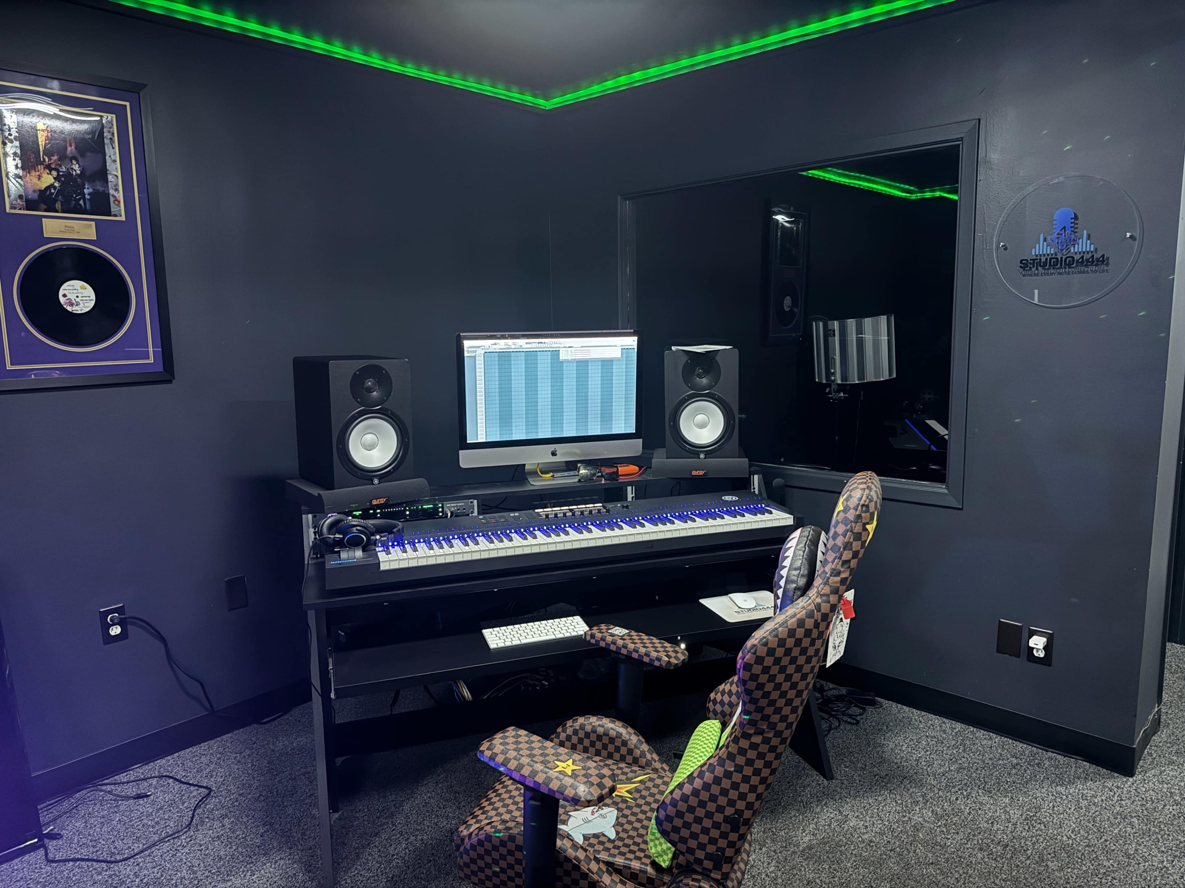The image shows a music production studio with a computer setup, speakers, and a keyboard on a desk, surrounded by dark walls and green LED lighting.