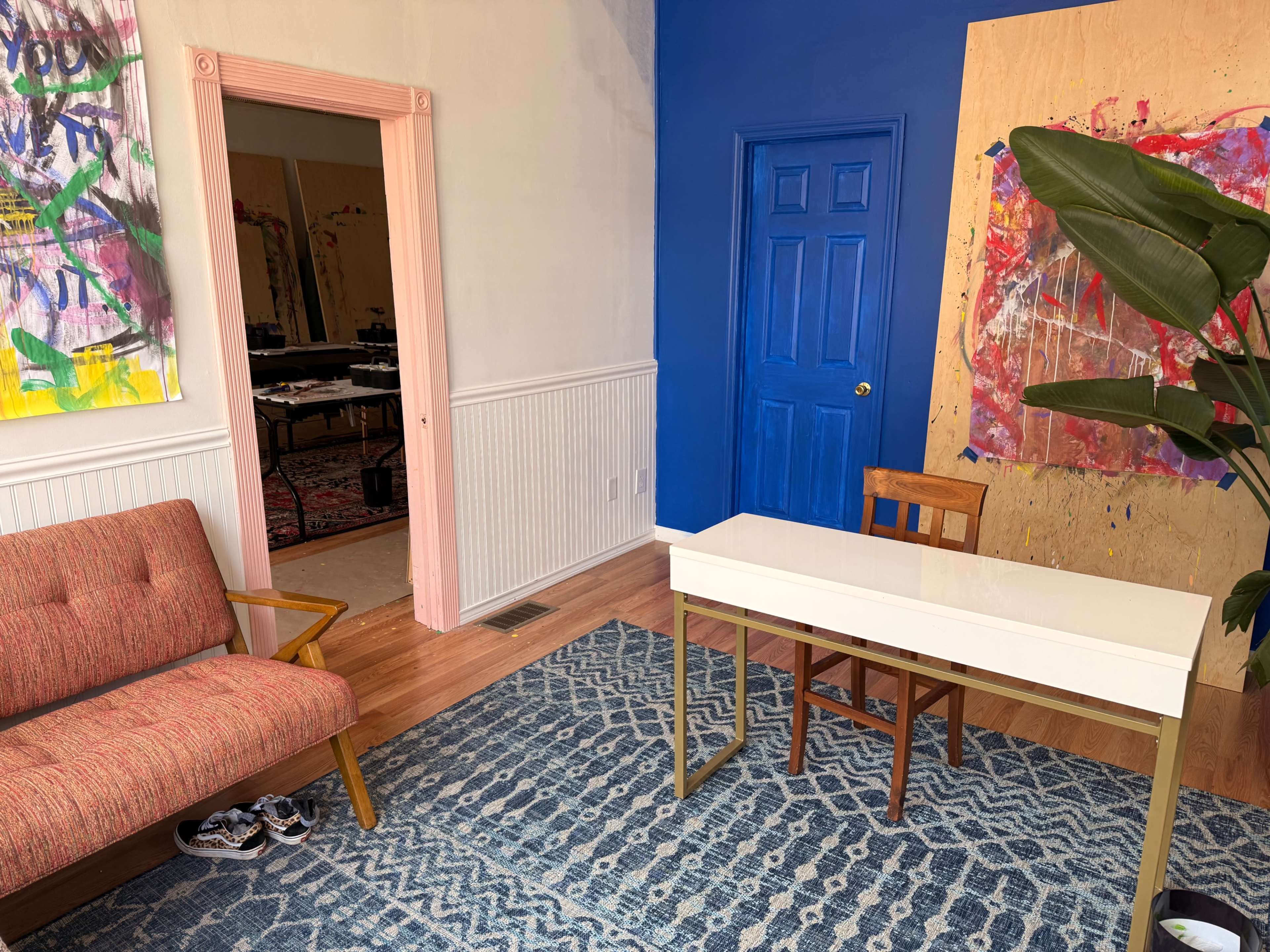 The image shows a modern, bright room featuring a white desk with a wooden chair, a pink-framed doorway, a colorful abstract painting on a blue wall, and a patterned rug.