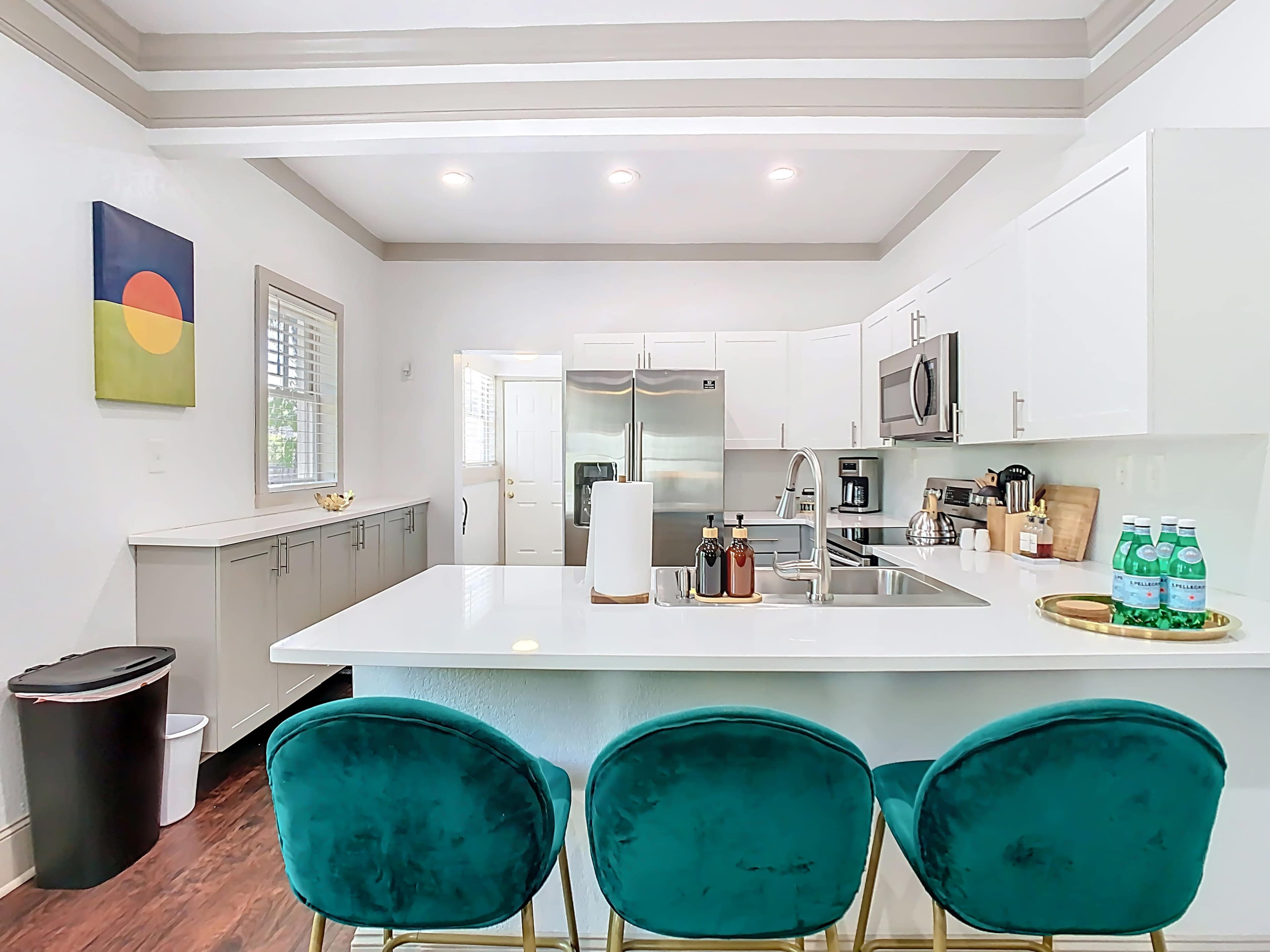 The image shows a modern kitchen with white cabinetry, stainless steel appliances, and a central island with teal bar stools.