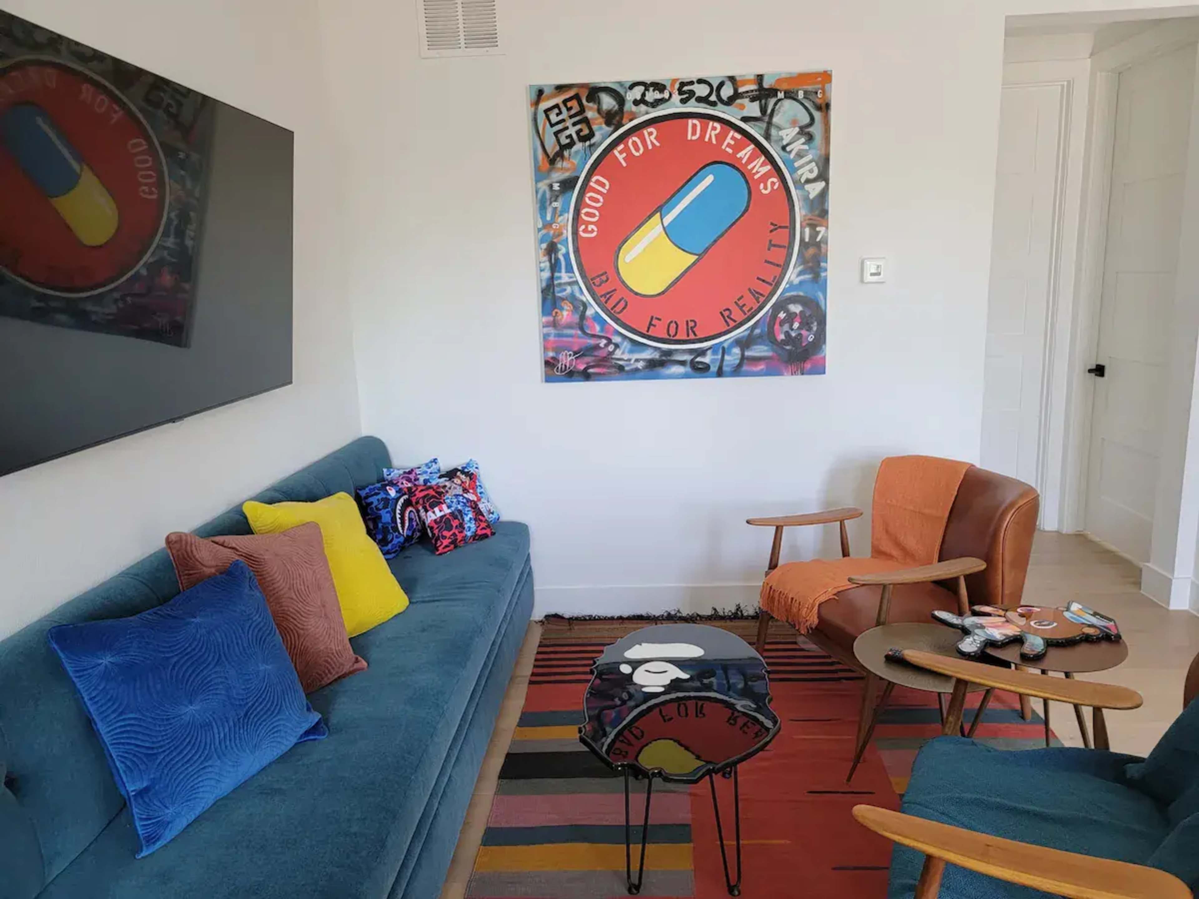 A living room features a blue sofa with colorful pillows, an orange chair, and a wall adorned with a large artwork featuring a pill design.