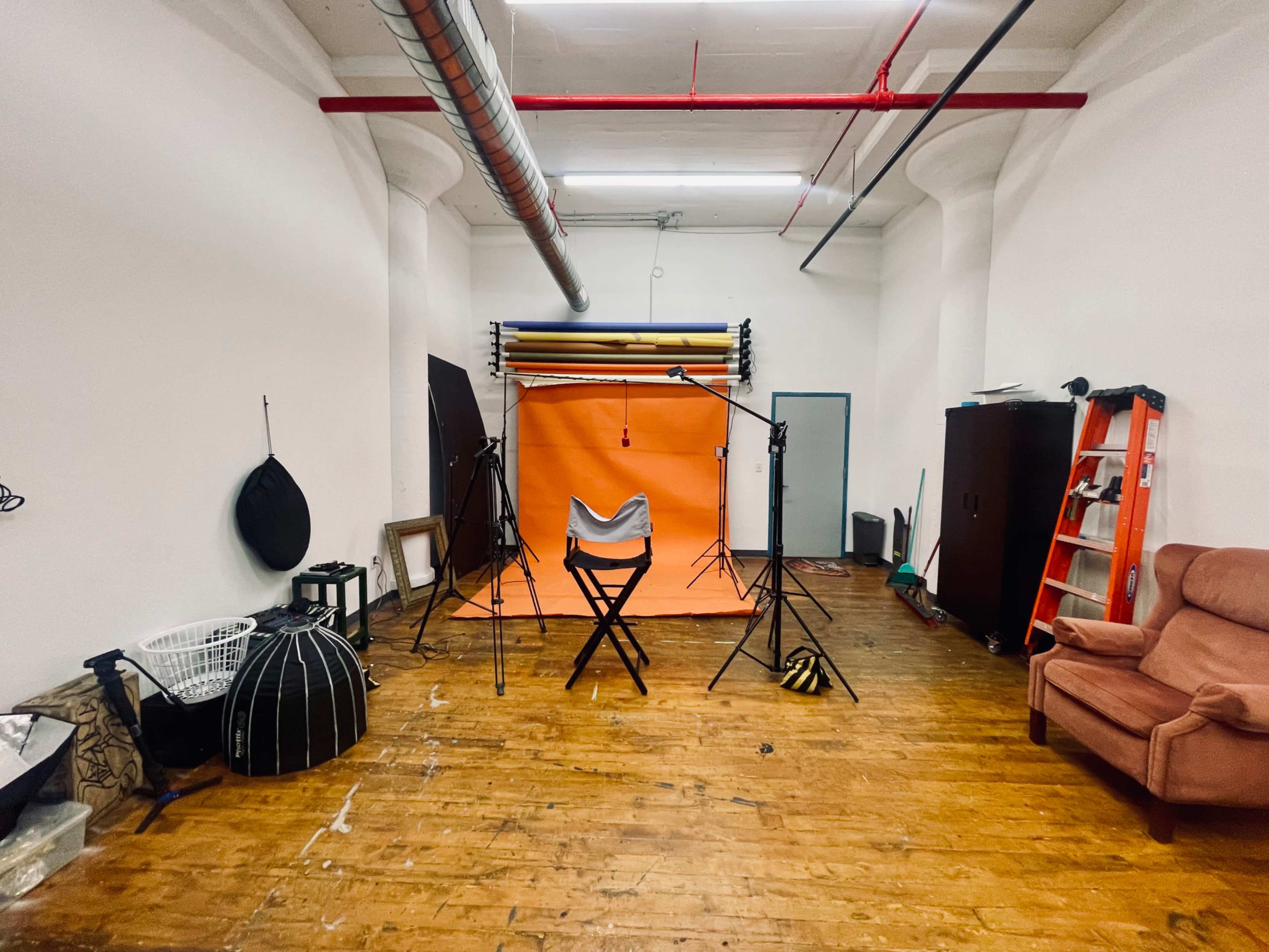 The image shows a photography studio featuring an orange backdrop, various lighting equipment, a folding chair, and a brown sofa, all set in a room with wooden flooring.