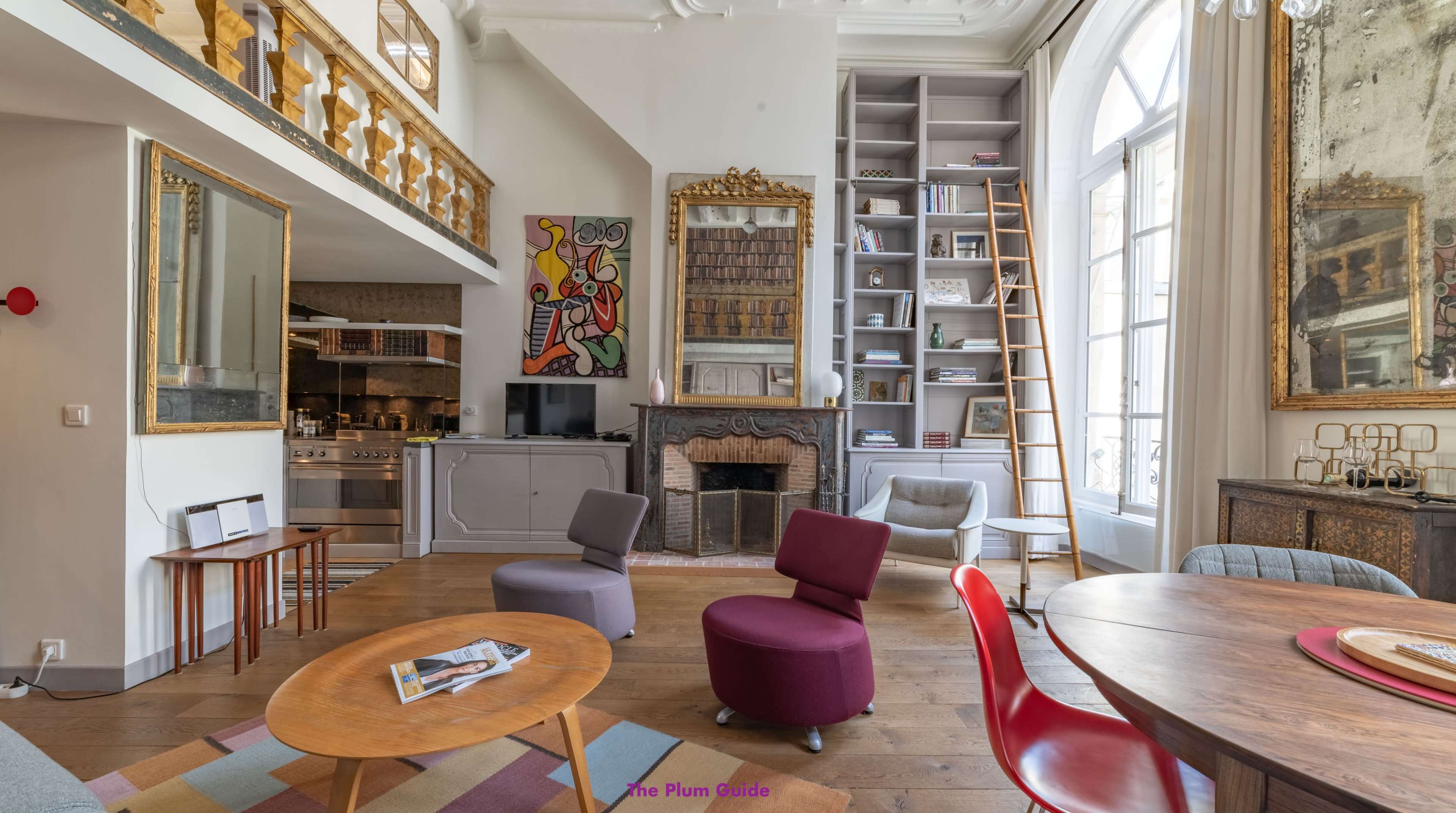 A cozy, well-decorated living space featuring a wooden floor, a fireplace, a circular dining table, colorful chairs, and large windows with natural light.