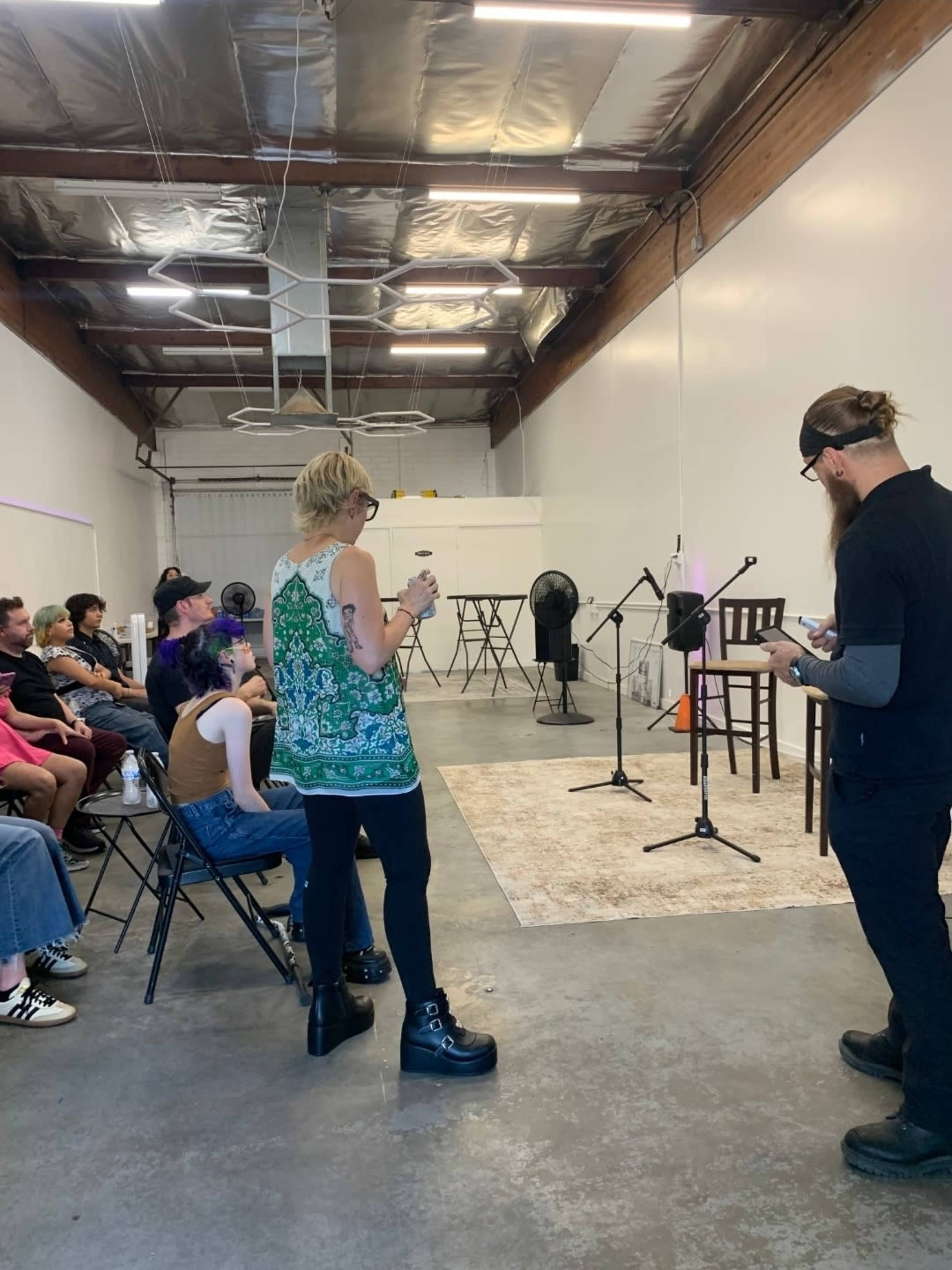 A group of people is seated in folding chairs, facing a small stage with two microphones and chairs in a spacious, industrial-style room.