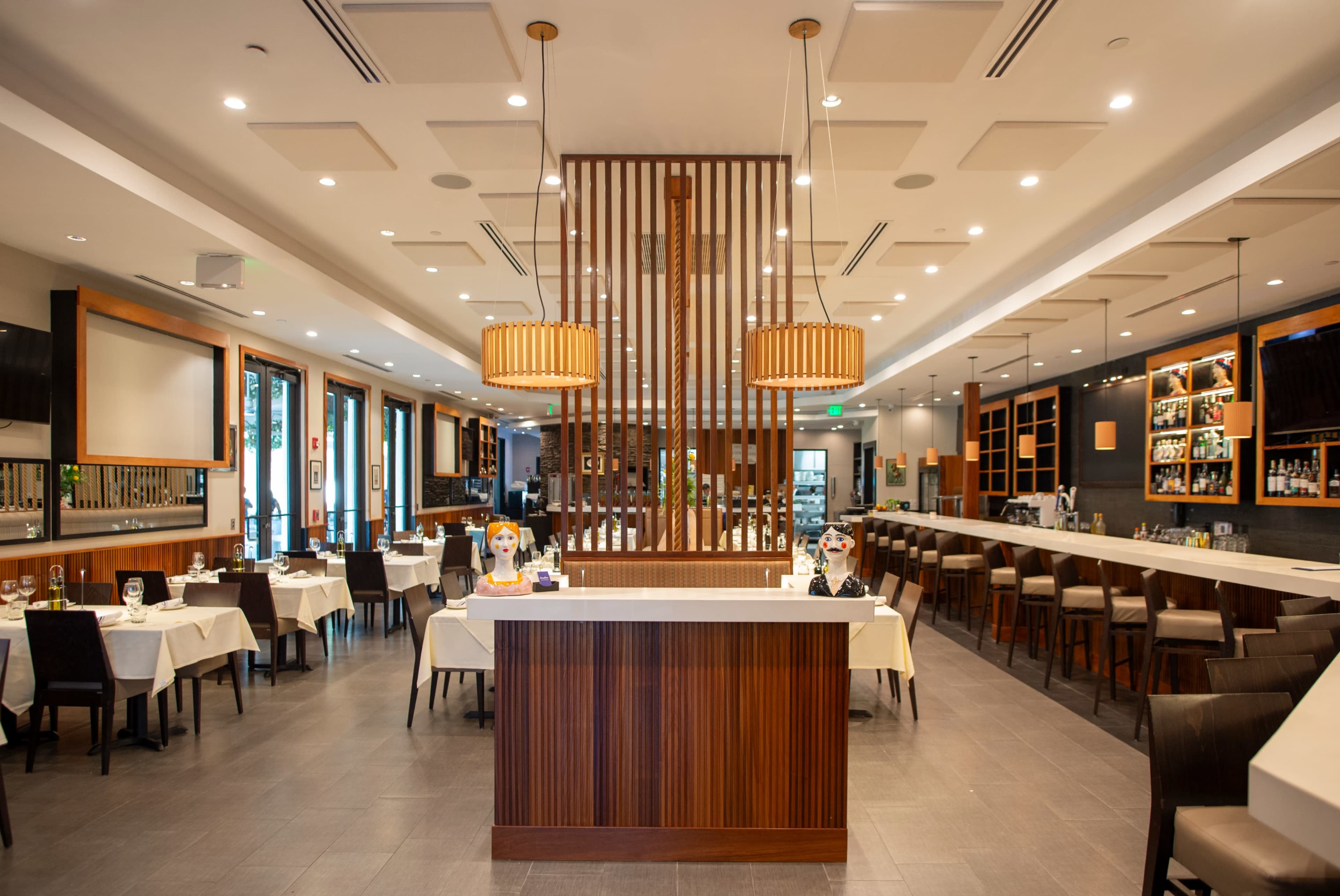 The image shows a modern restaurant interior featuring a central reception area, dining tables with white tablecloths, and a bar counter in the background.