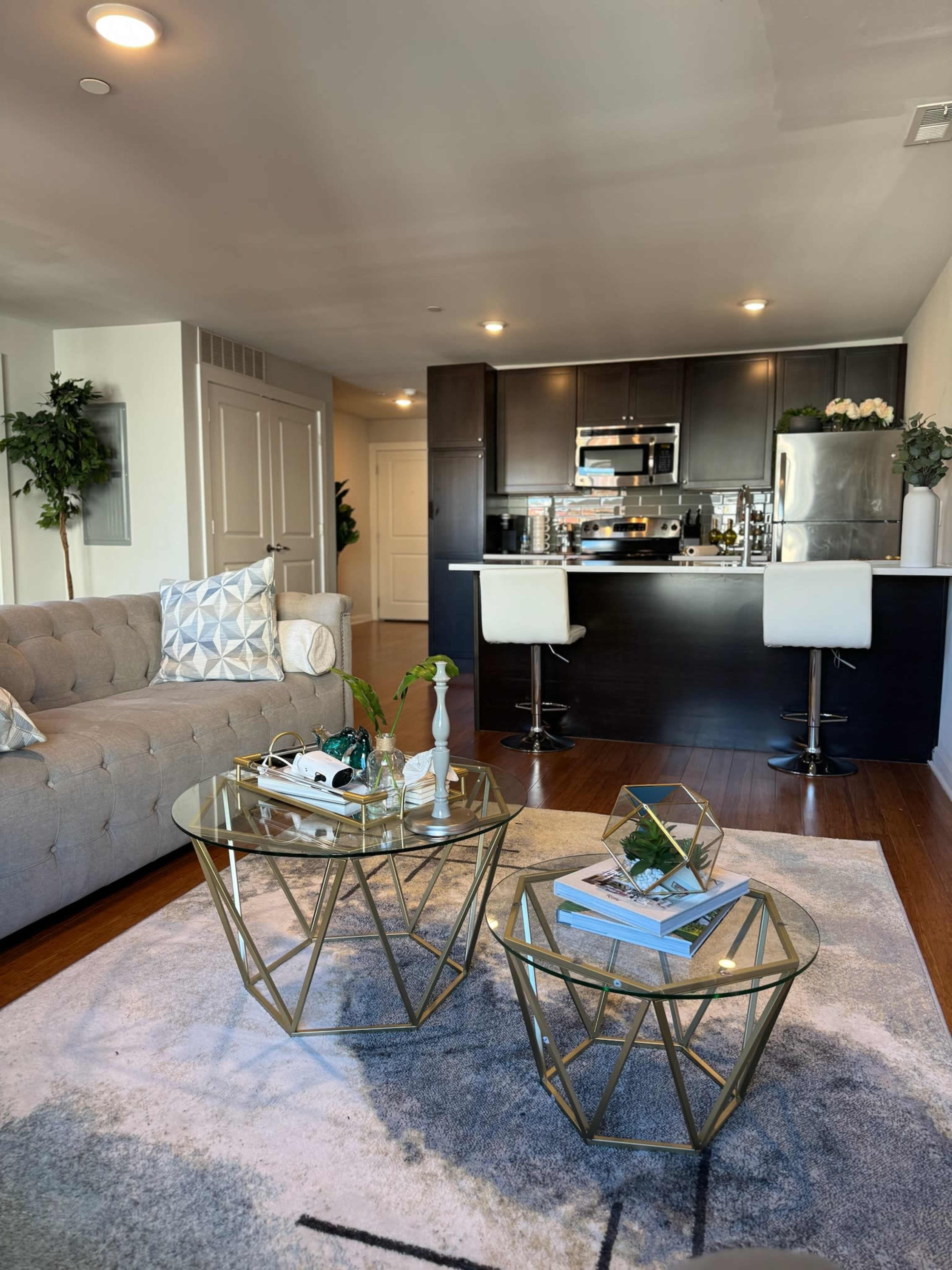 The image shows a modern living room with a beige sofa, two glass-top coffee tables, and an open kitchen in the background featuring dark cabinetry and bar stools.
