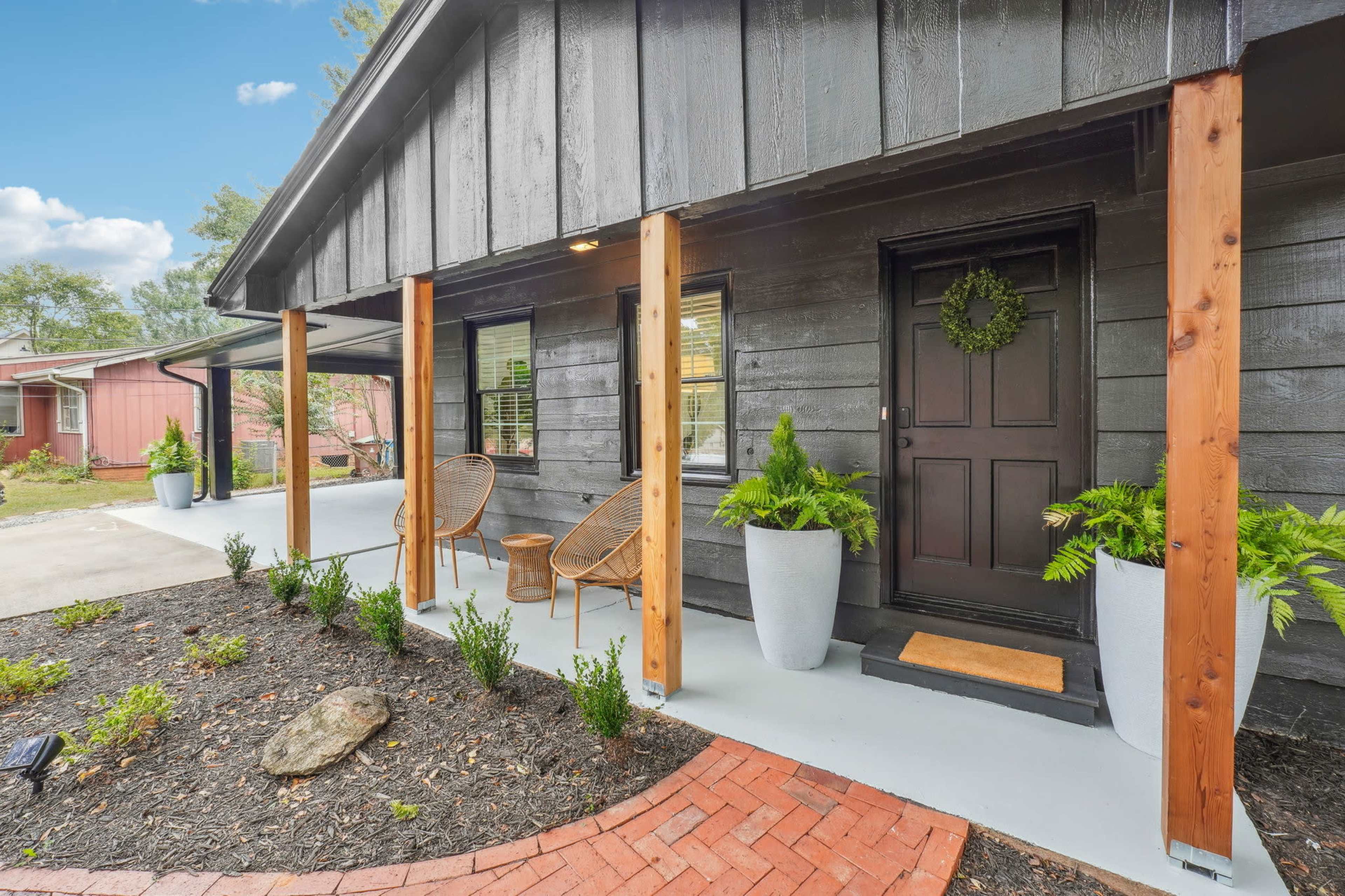 A front porch of a house featuring two chairs, a welcome mat, and large planters with greenery, surrounded by a brick walkway and mulch.