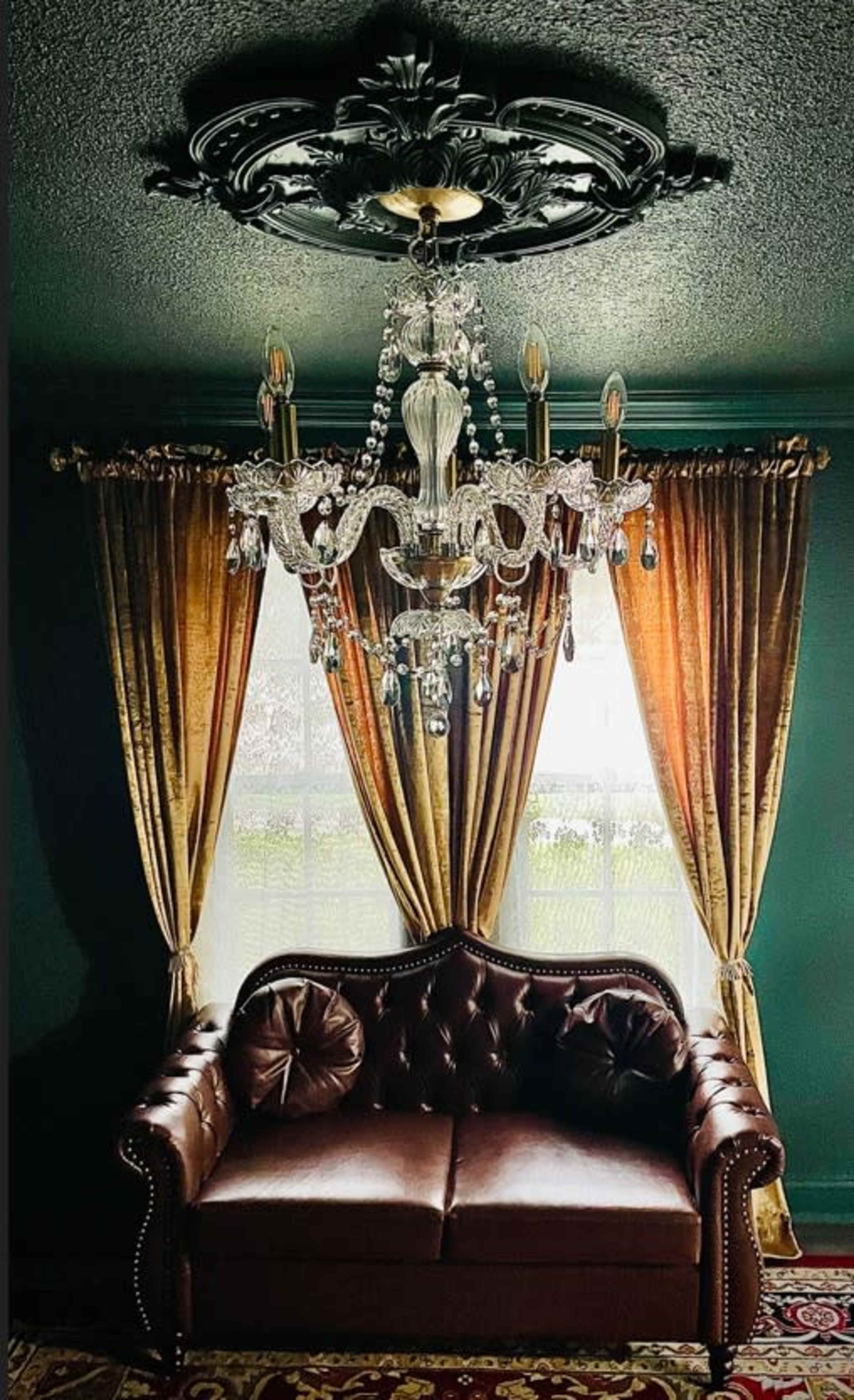 The image features a brown leather sofa beneath an ornate chandelier, with elegant golden curtains framing a window in a richly colored room.