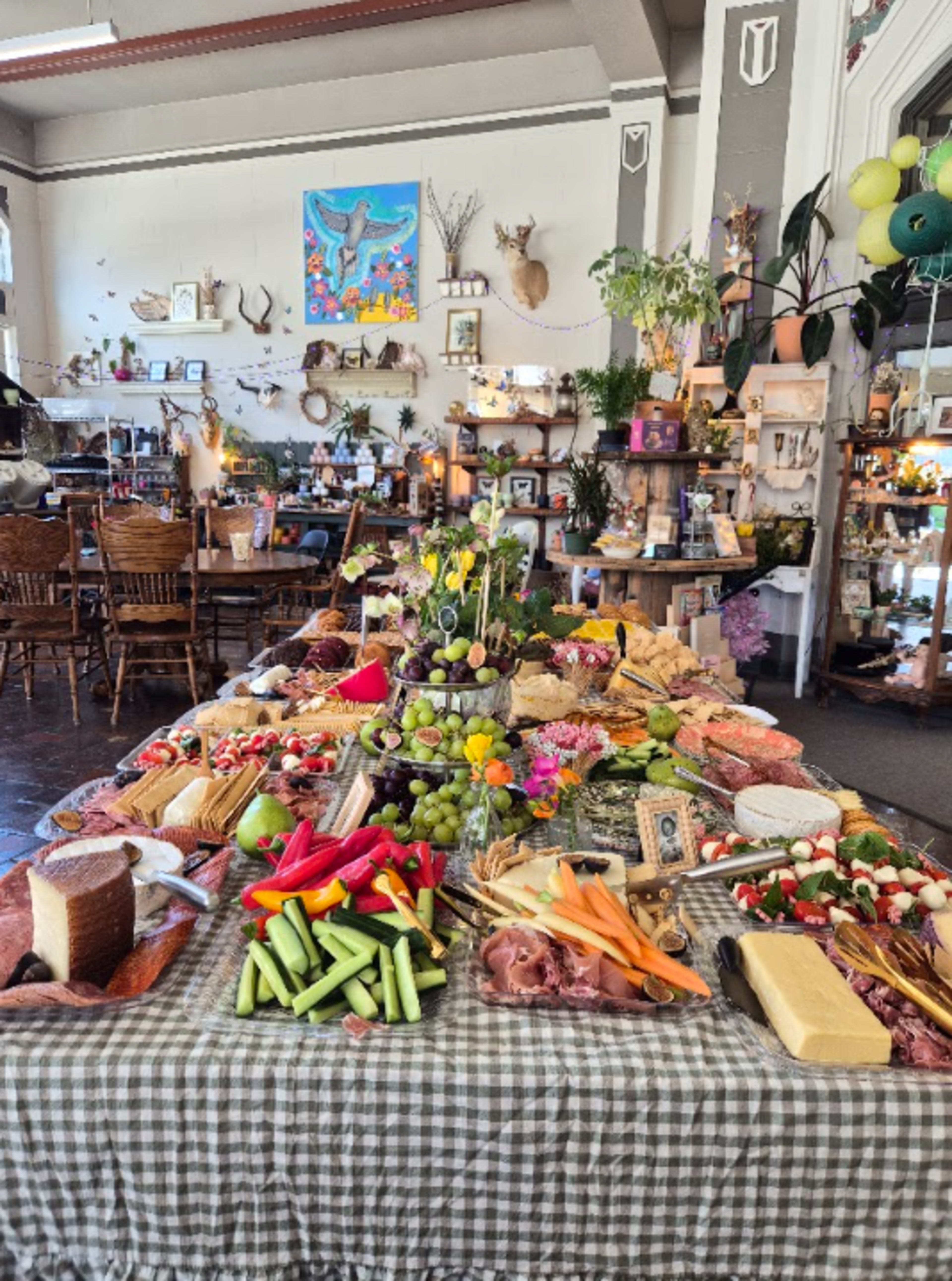 A long table is covered with a variety of fresh fruits, vegetables, cheeses, and meats, arranged on a checkered tablecloth in a decorated indoor space.