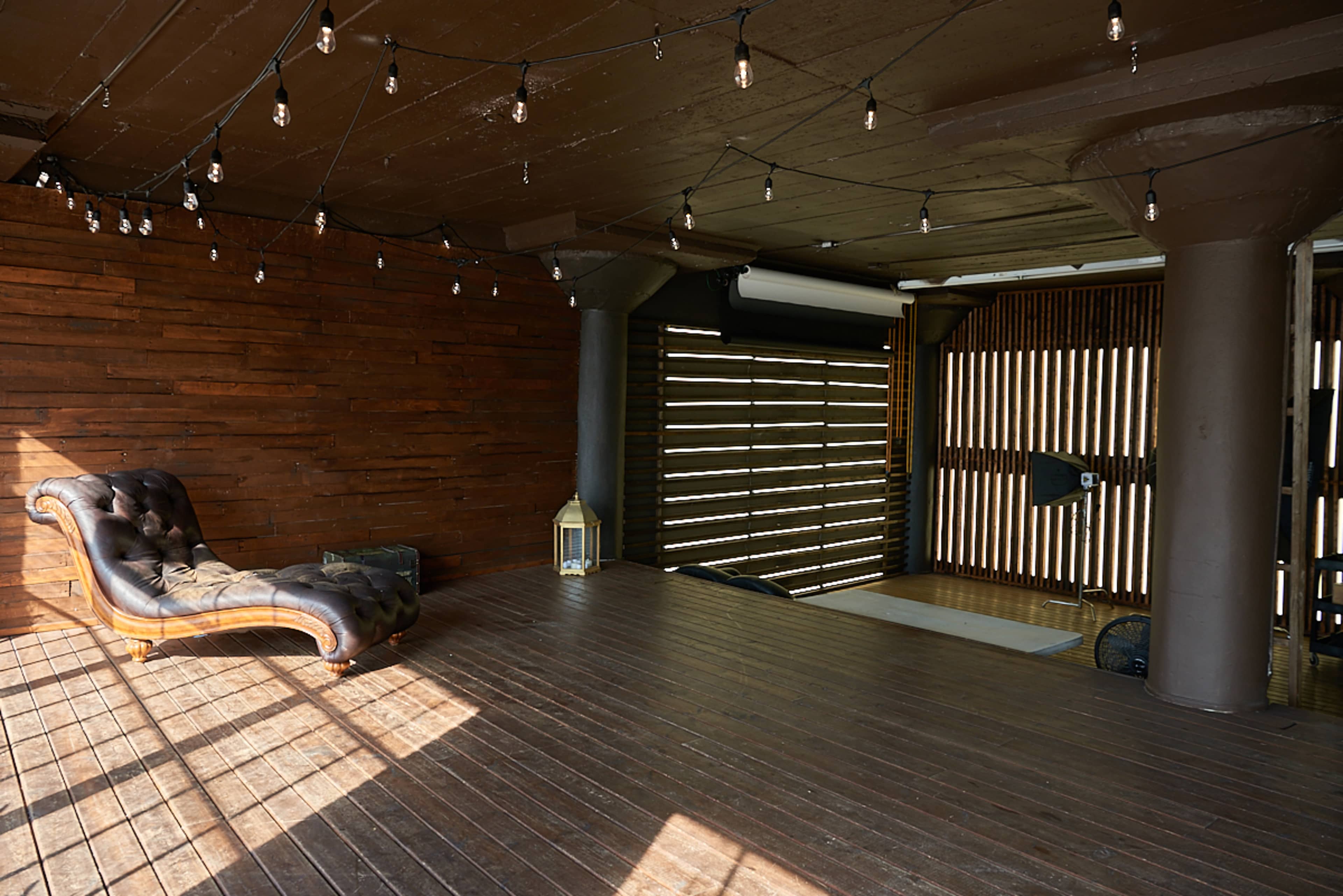 A spacious room with wooden walls and flooring, featuring a chaise lounge and string lights overhead.