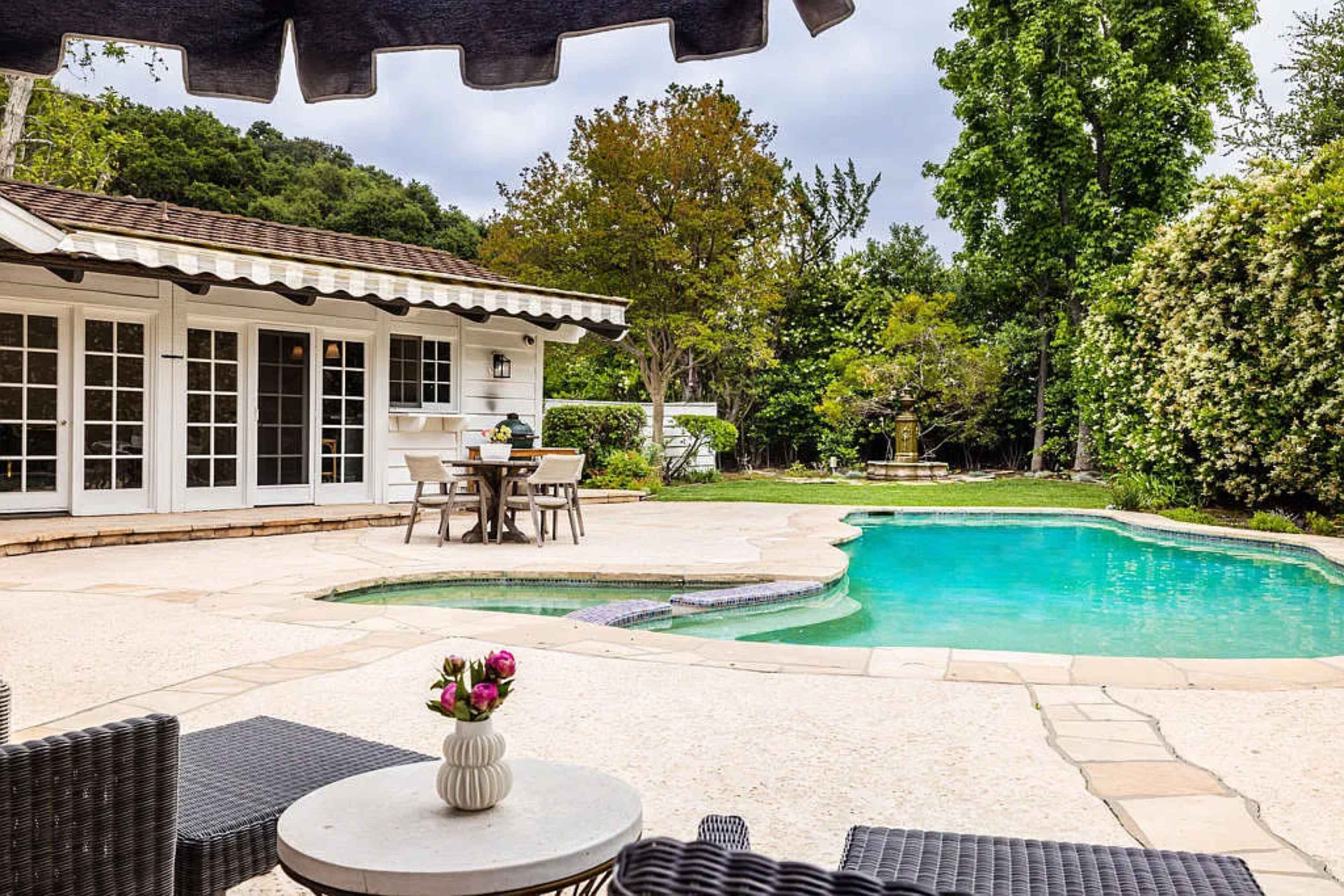 A backyard features a swimming pool surrounded by a patio, dining table, and lush greenery.
