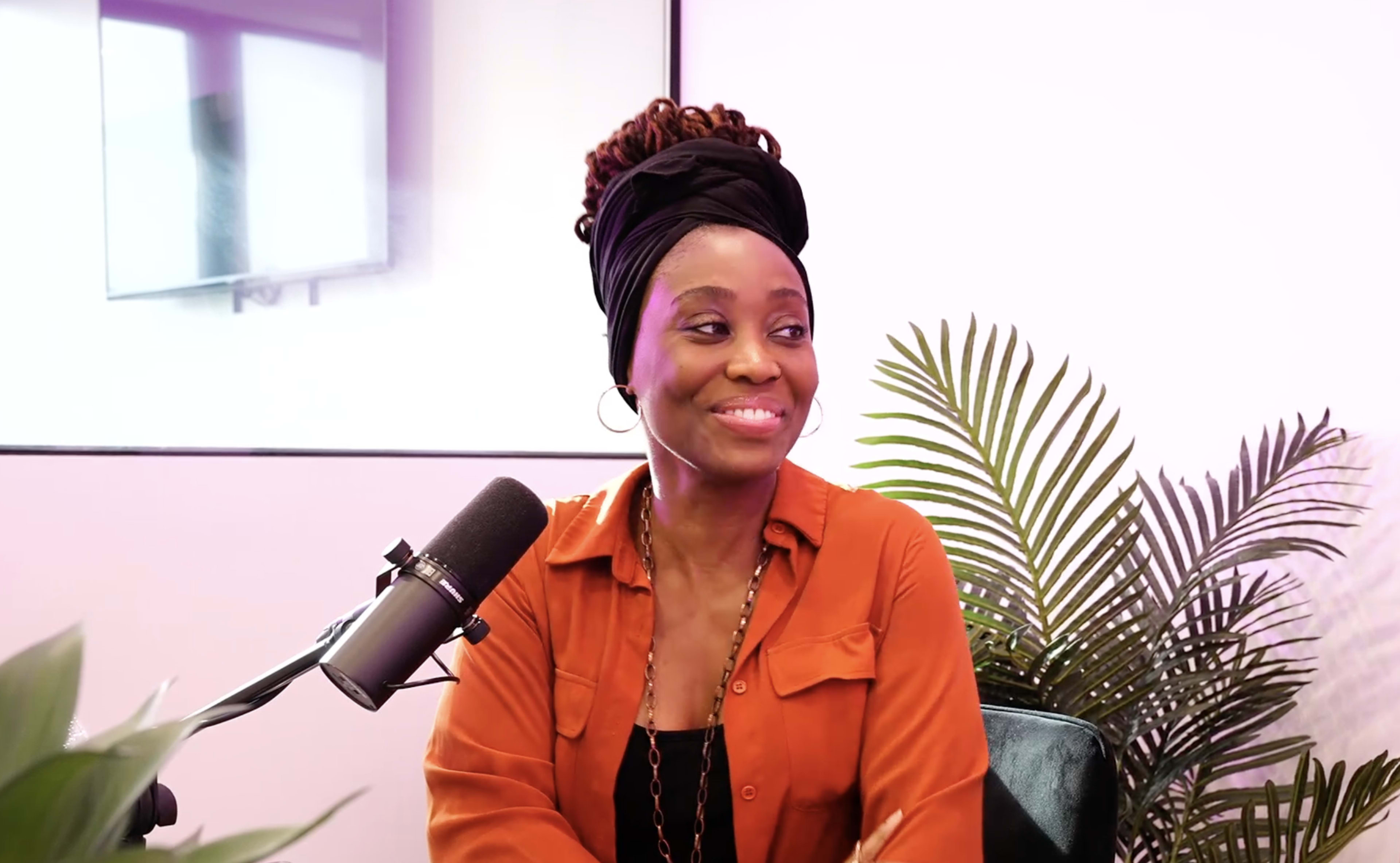 A woman with a head wrap and orange shirt sits in front of a microphone and a plant.
