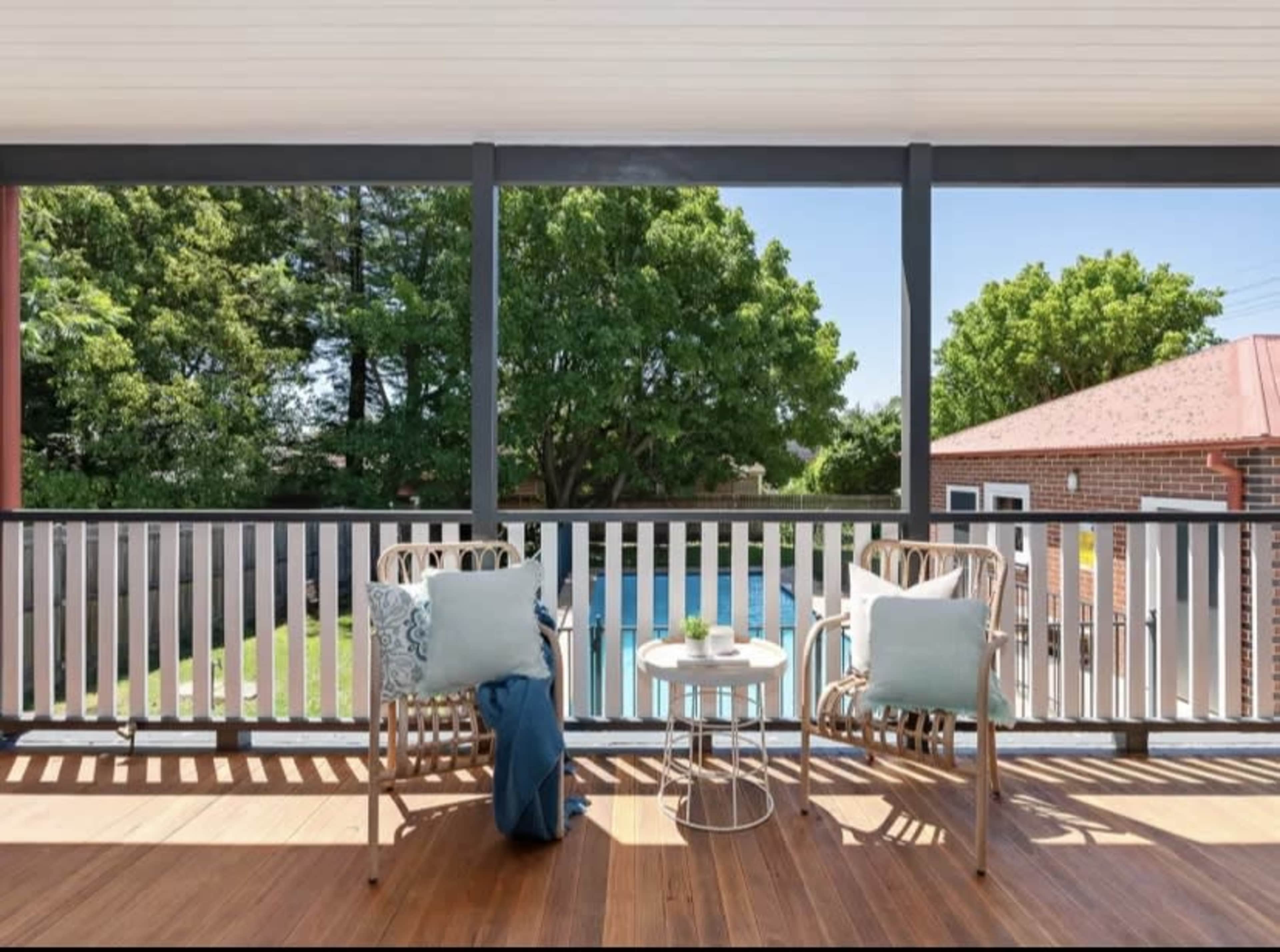 A covered balcony with two chairs and a small table, overlooking a backyard pool surrounded by greenery.