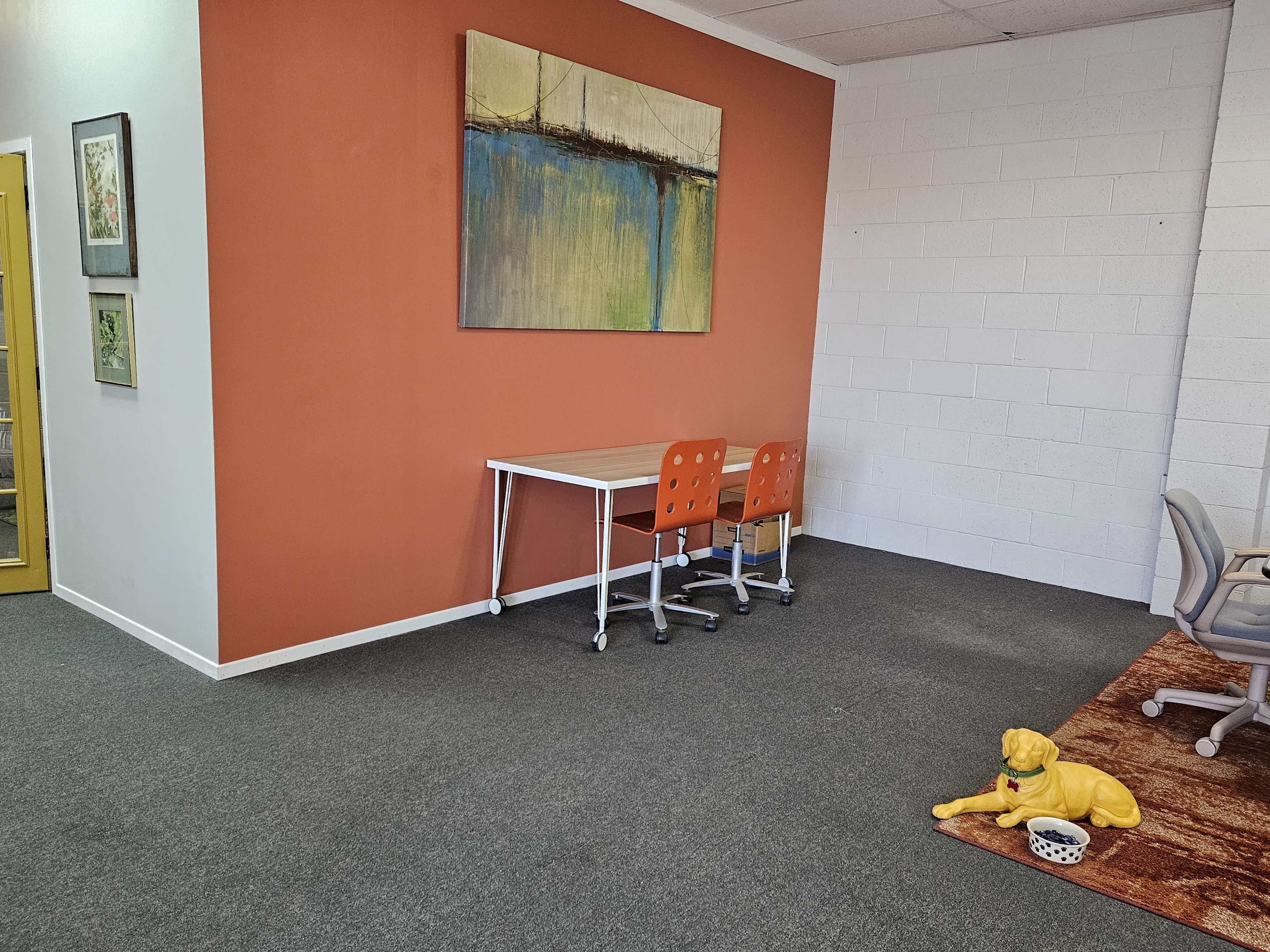 A modern office space featuring an orange wall, a small table with orange chairs, and a yellow dog statue on the floor.