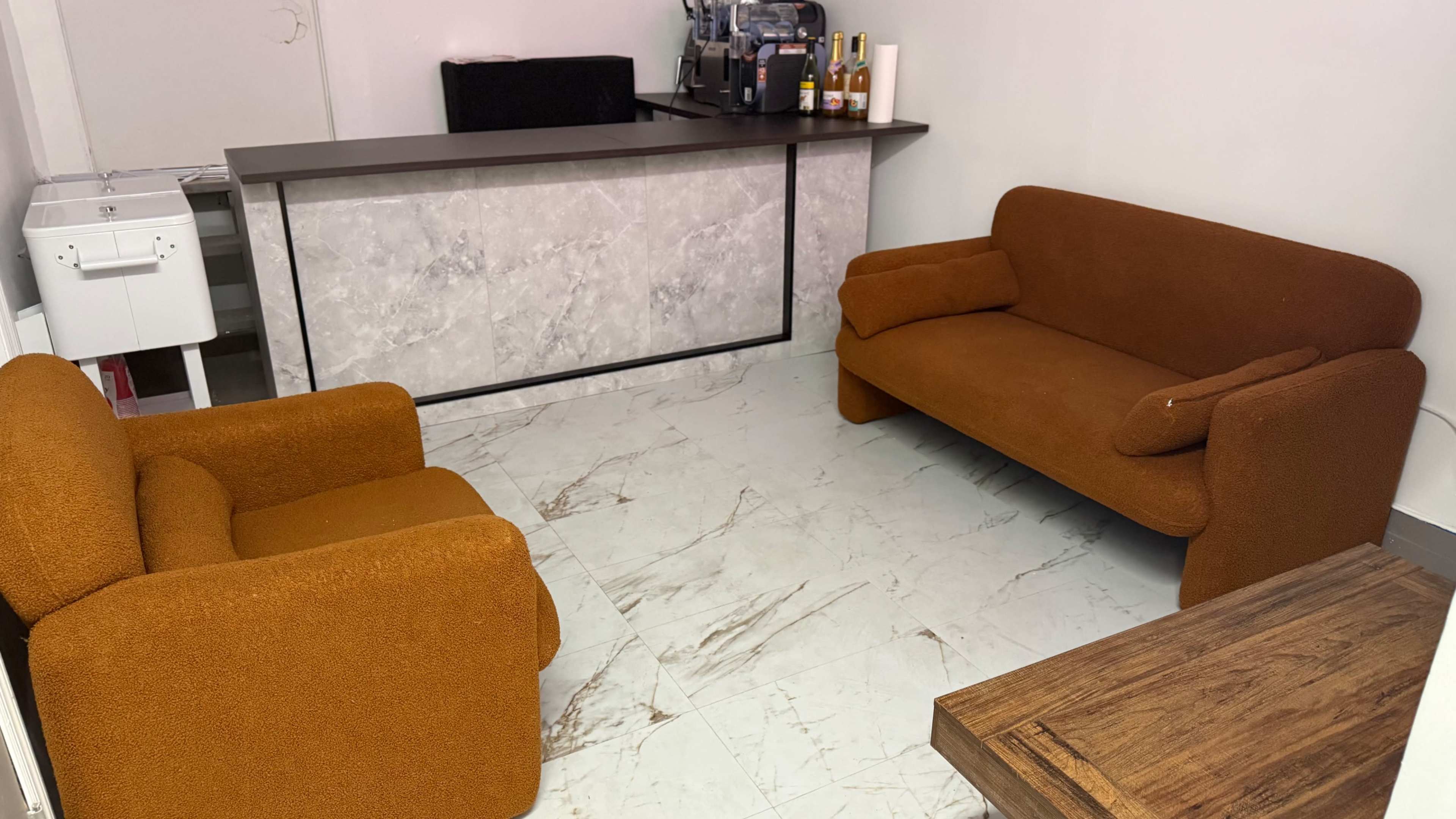 The image shows a small waiting area with two orange sofas and a reception counter made of marble-like material.