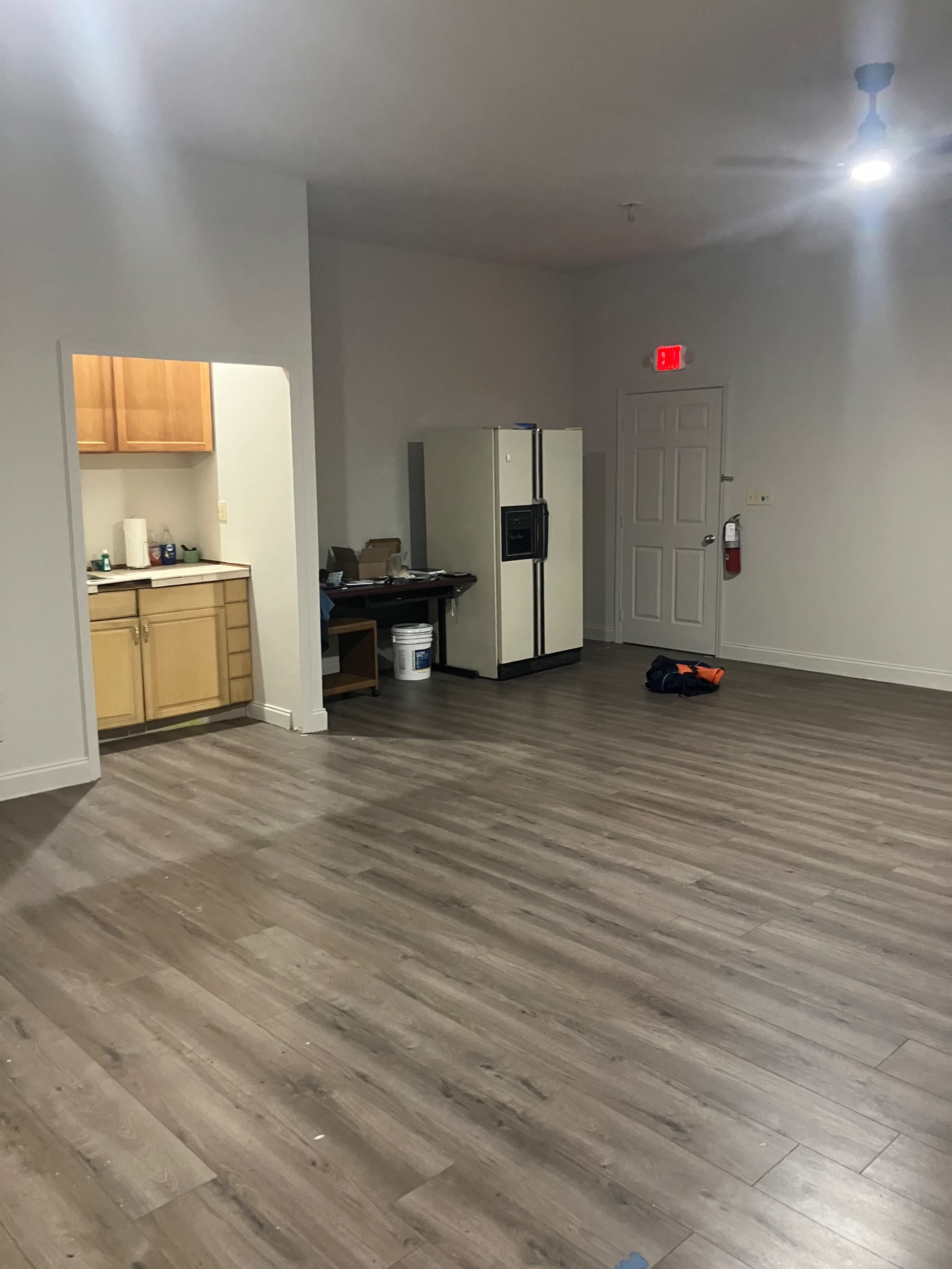 The image shows a spacious room with wooden flooring, a kitchen area on the left featuring light wood cabinetry and a white refrigerator, and an open space that appears to be empty except for a few items on the floor.
