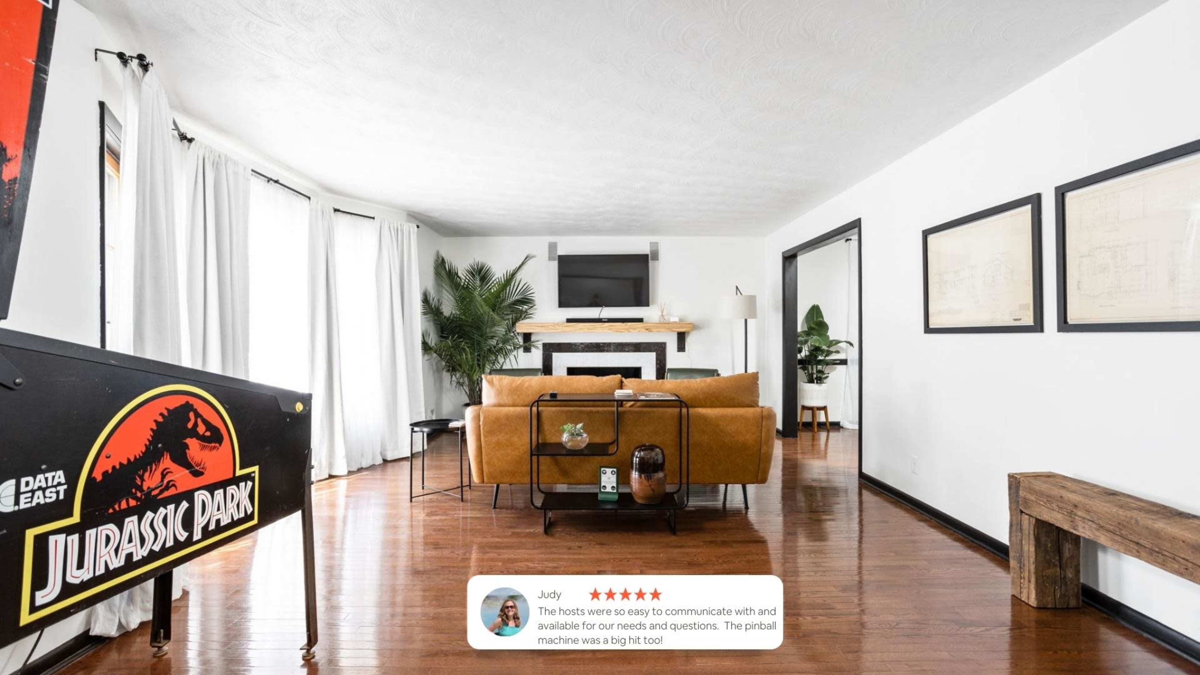 The scene shows a modern living room with hardwood floors, a large brown sofa, a television mounted above a white mantel, and a Jurassic Park sign in the foreground.