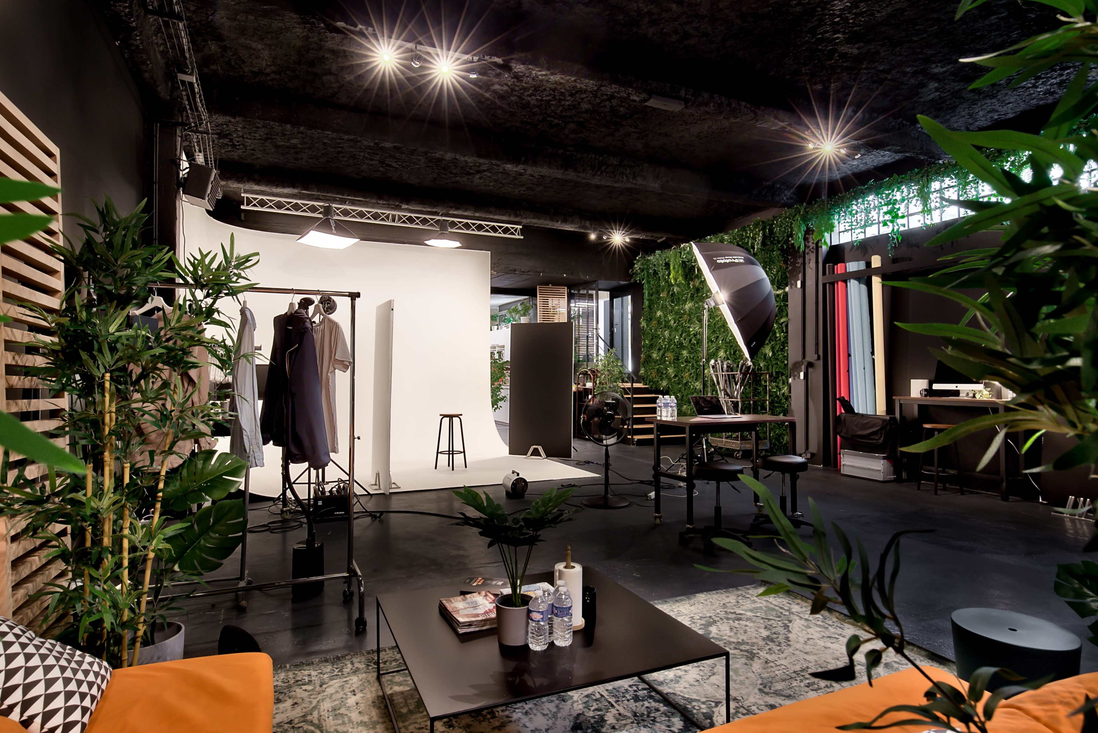 A photography studio featuring a white backdrop, various lighting equipment, and green plants alongside furniture like a table and chairs.