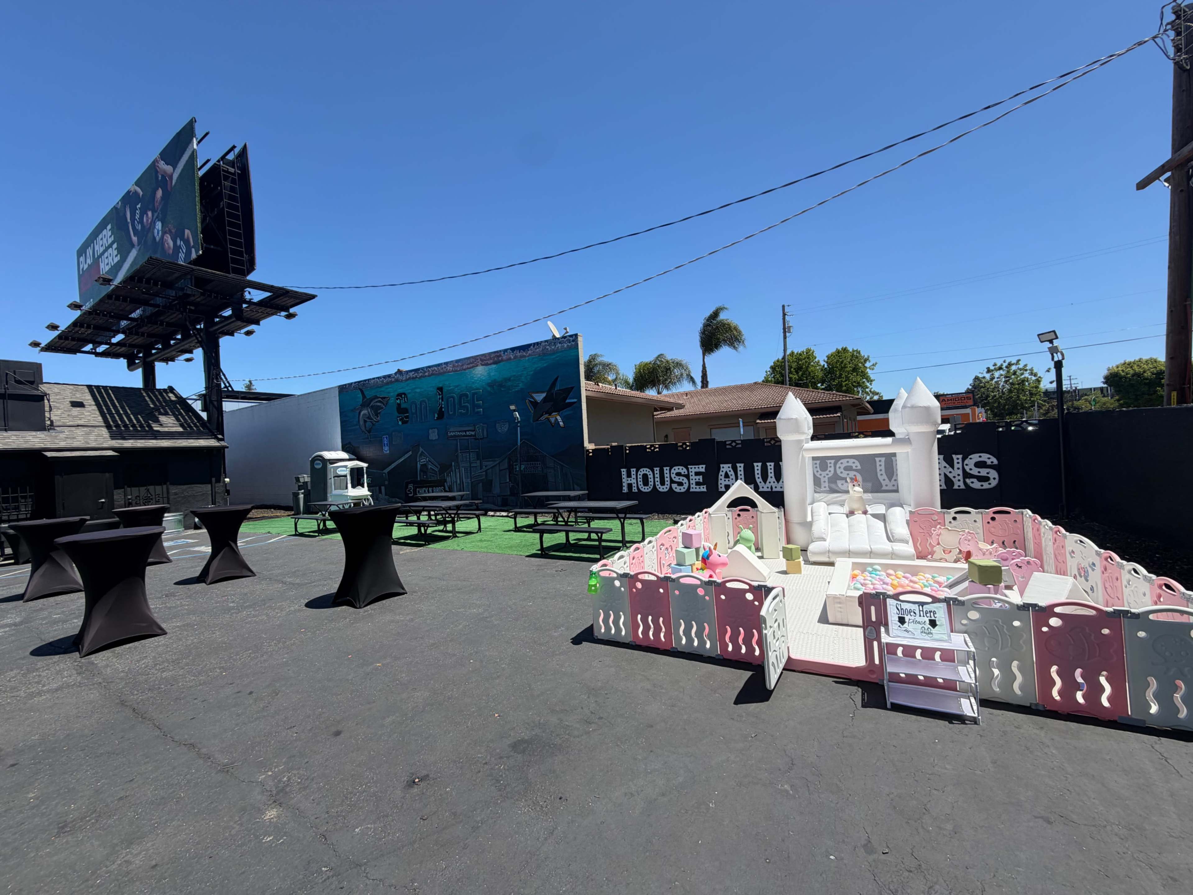 The image shows an outdoor venue featuring a fenced play area with a small play structure, tables set up for gatherings, and a large billboard in the background.
