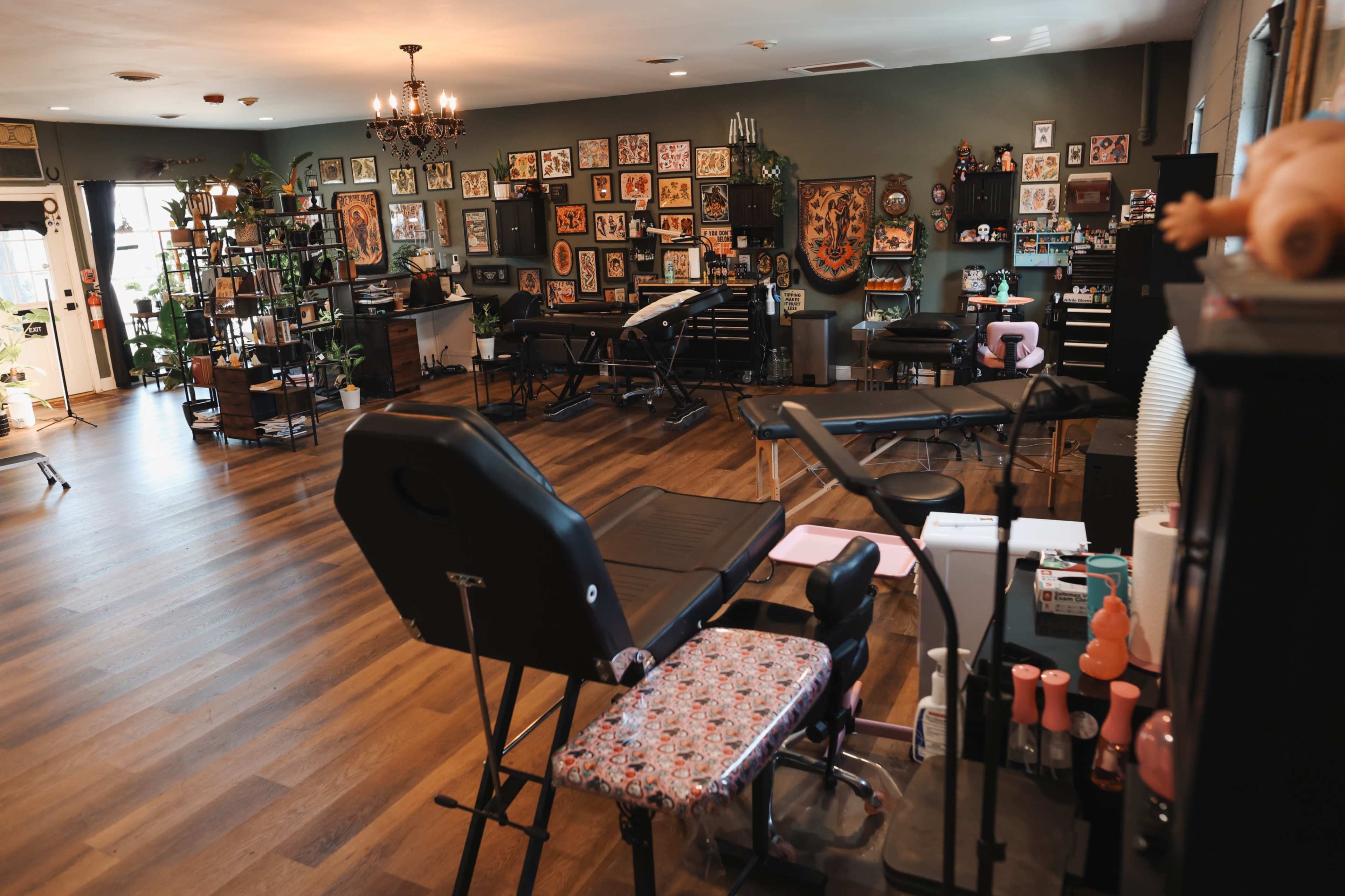 The image shows a spacious tattoo parlor with multiple workstations, tattoo equipment, and decorative artwork on the walls.