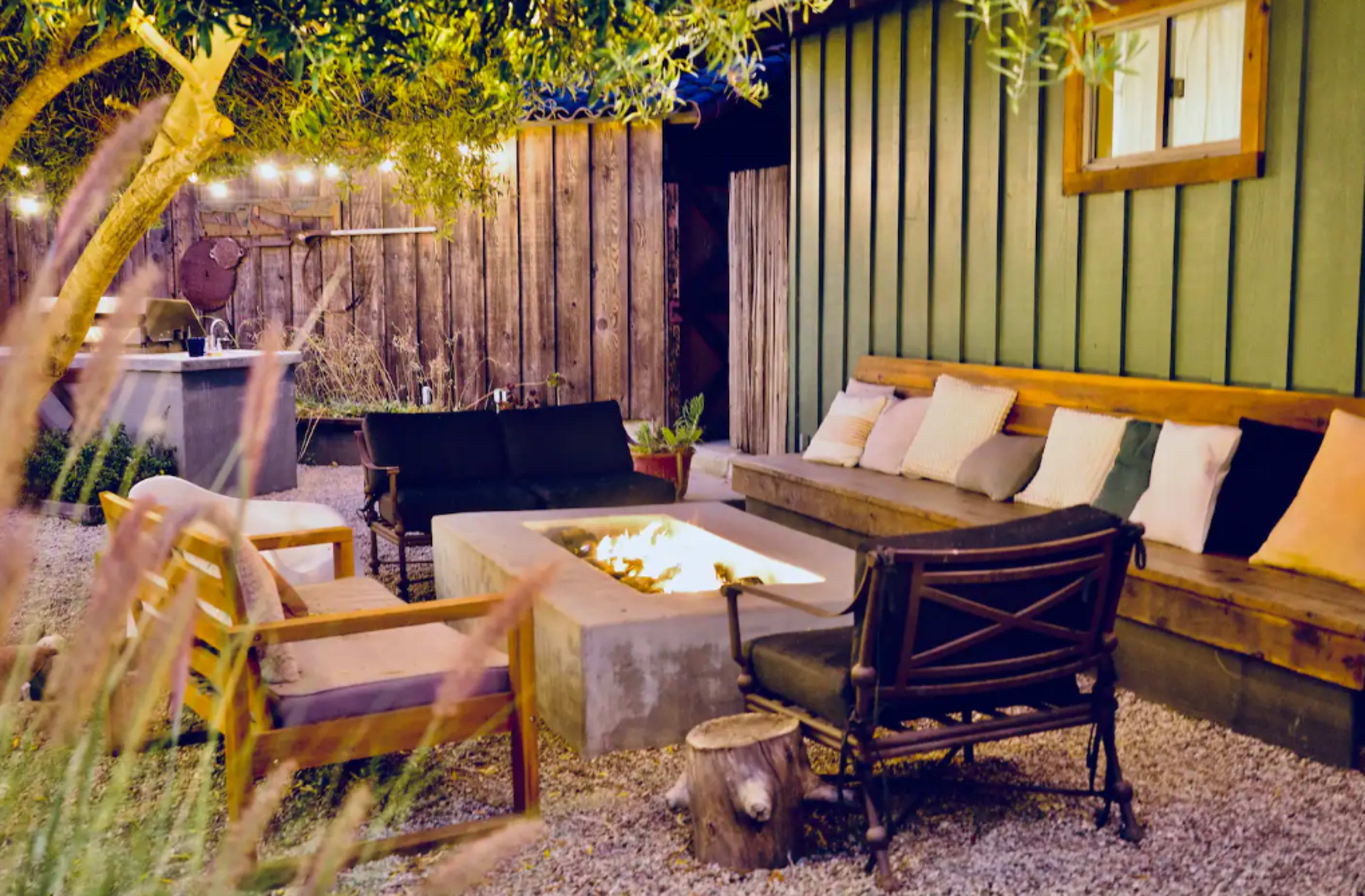 The image depicts a cozy outdoor seating area featuring a fire pit surrounded by various chairs and a wooden sofa against a green wooden wall.