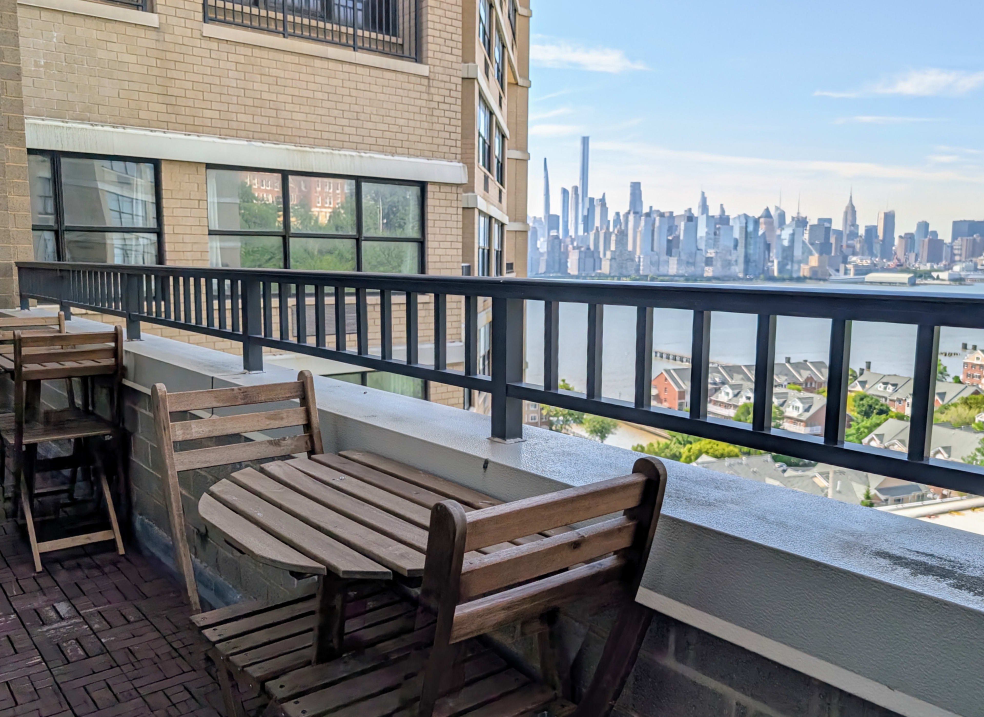 The image shows a balcony with wooden tables and chairs overlooking a river and a skyline of tall buildings.