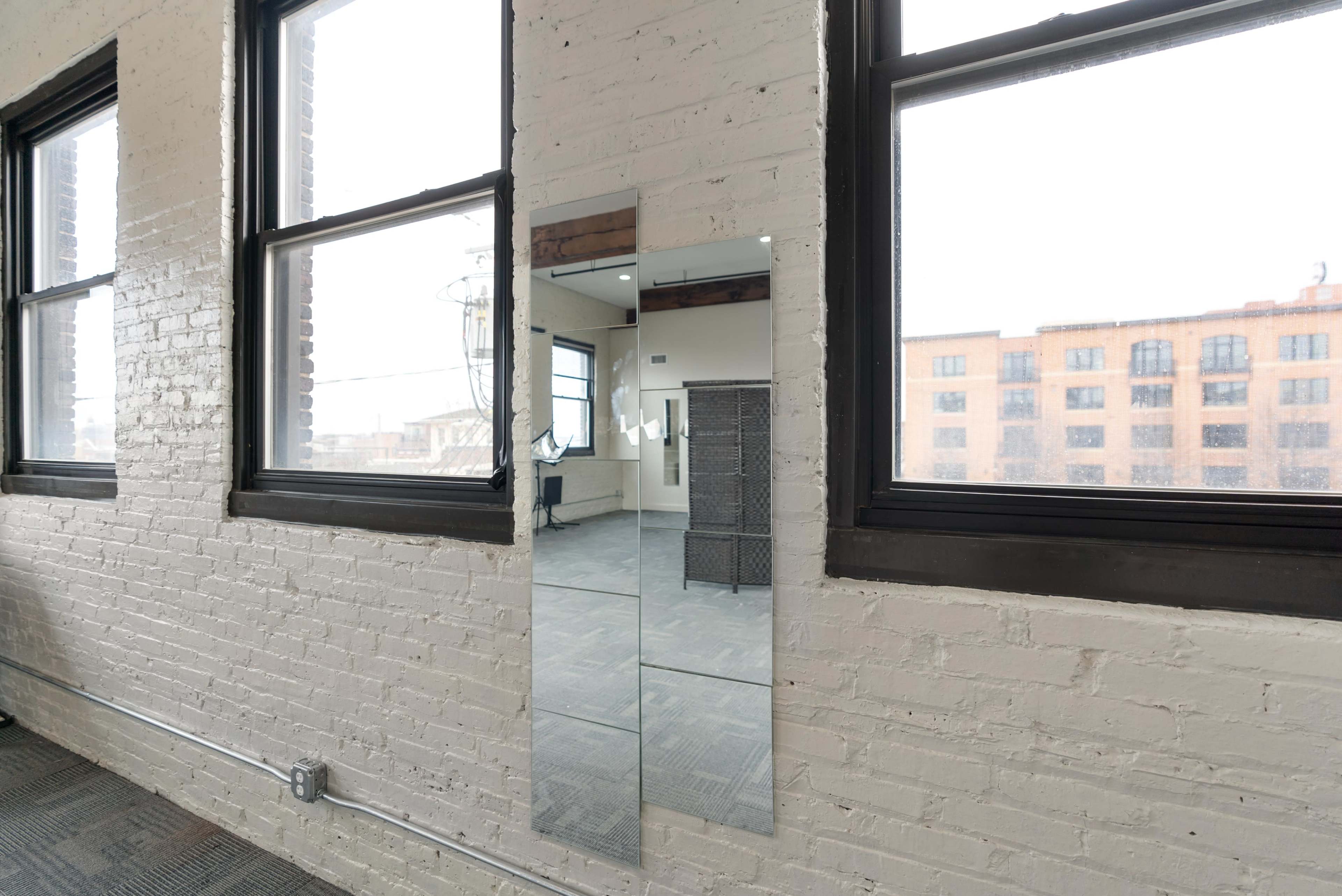 A rectangular mirror mounted on a white brick wall between two large windows in a room with gray carpet.