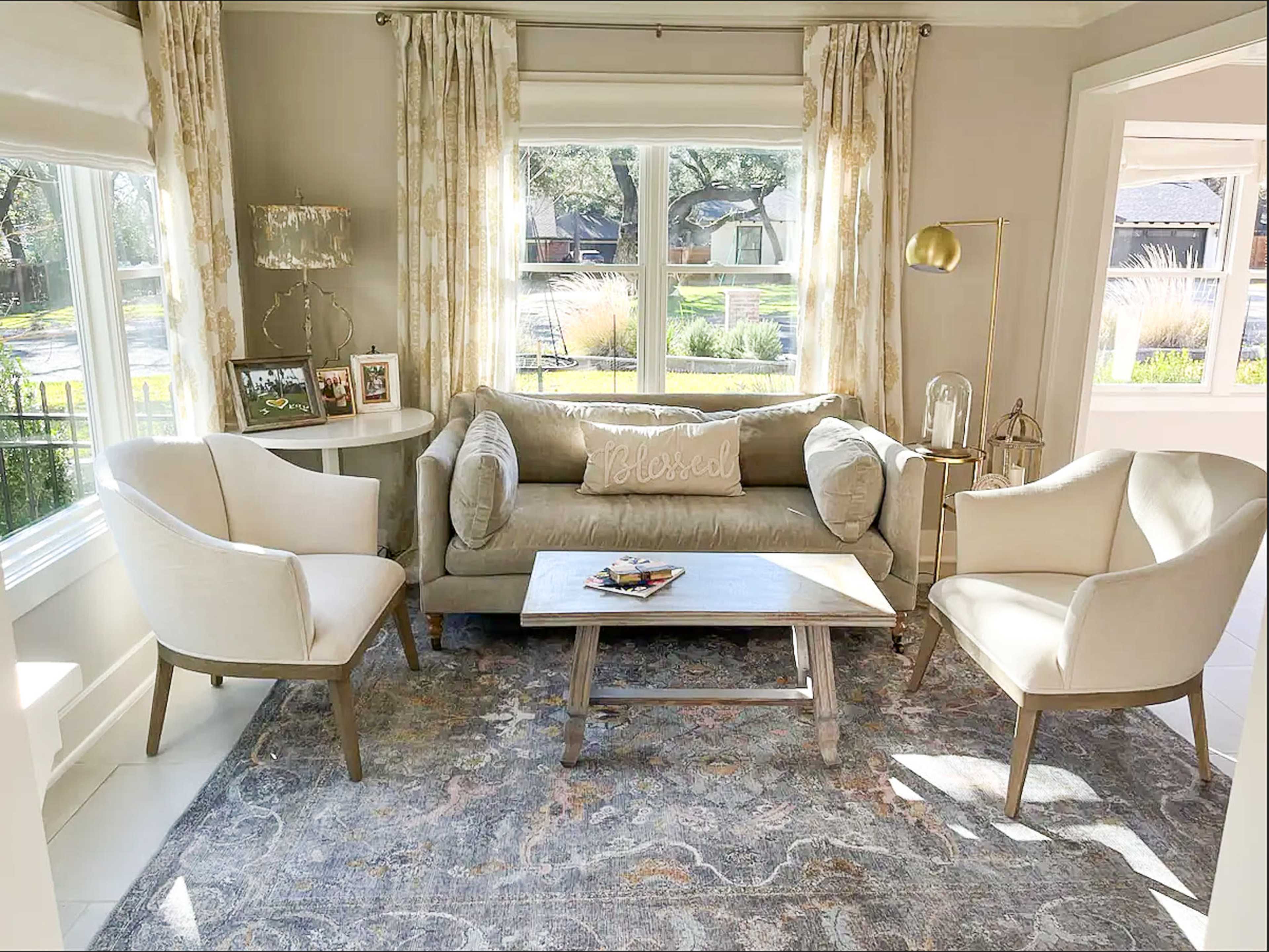 The scene shows a cozy living room with a beige sofa, two armchairs, and a coffee table on a patterned rug, all illuminated by natural light from large windows.