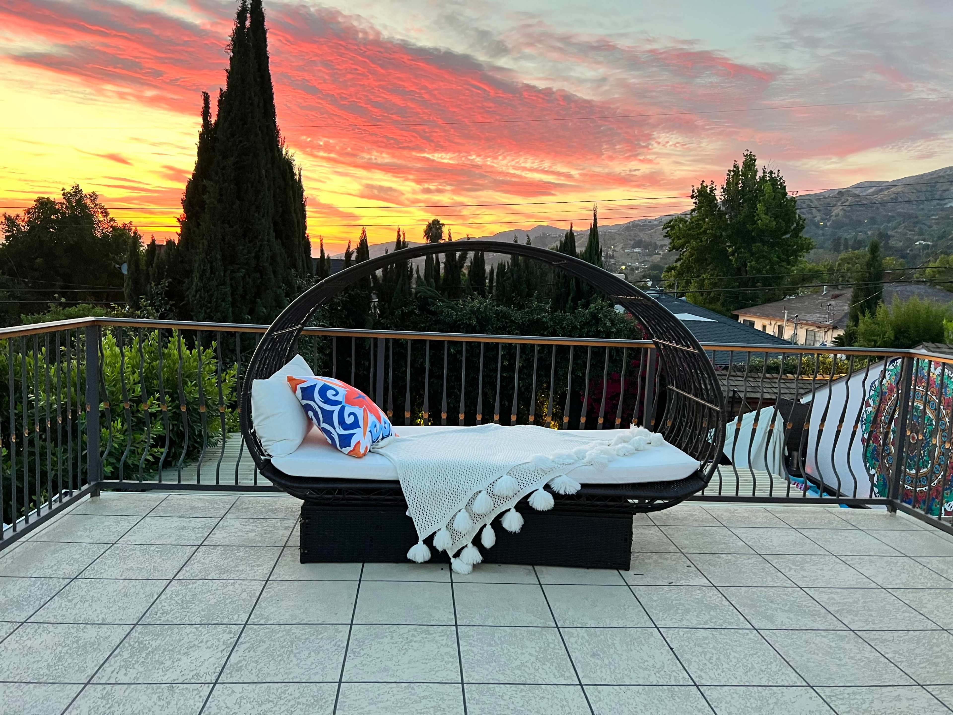 A wicker lounge chair with a white throw and decorative pillow sits on a tiled terrace, overlooking a vibrant sunset.