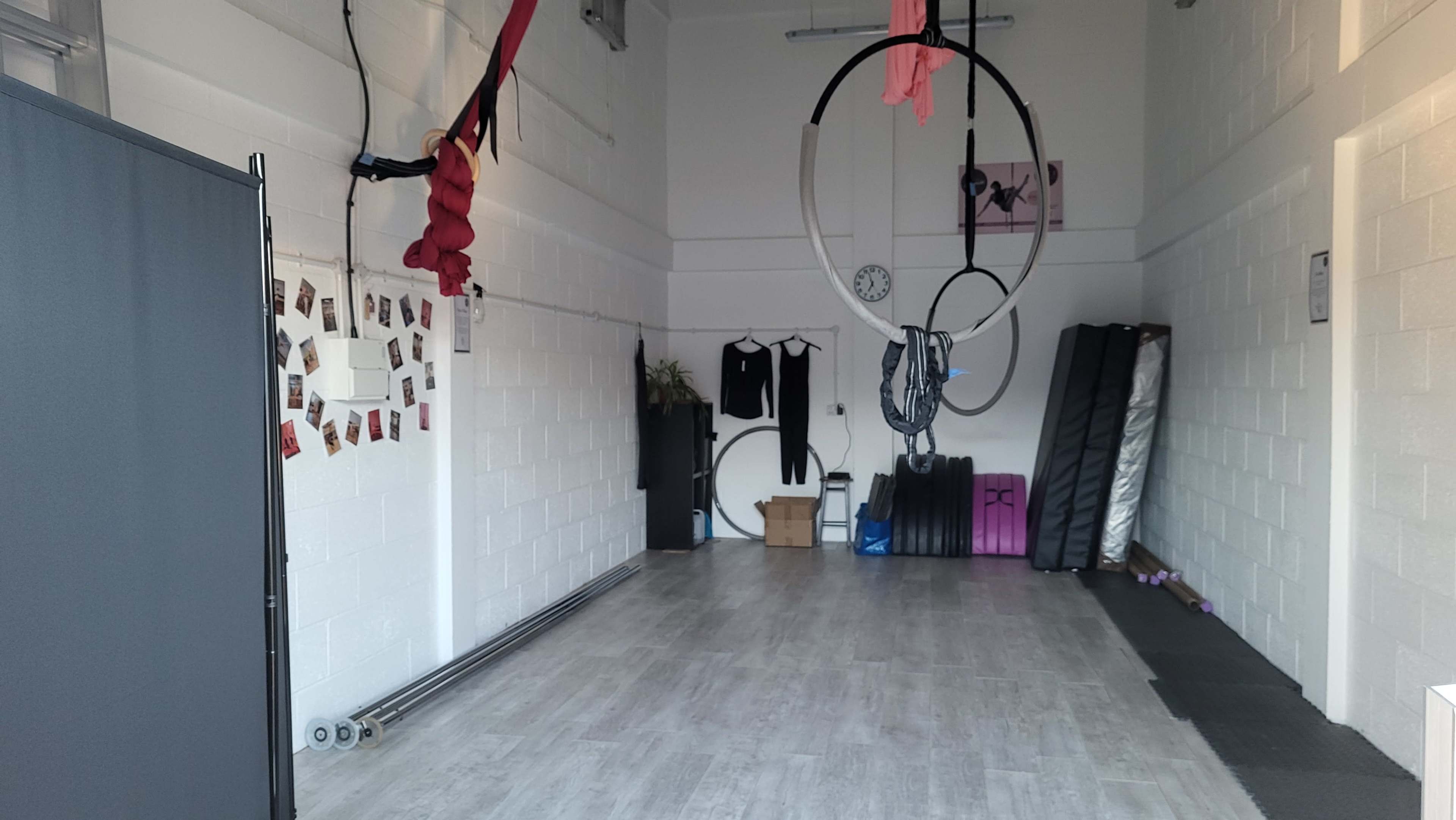 The image shows an empty room with white walls, featuring aerial silks, hoop apparatuses hanging from the ceiling, and fitness equipment stacked against one side.