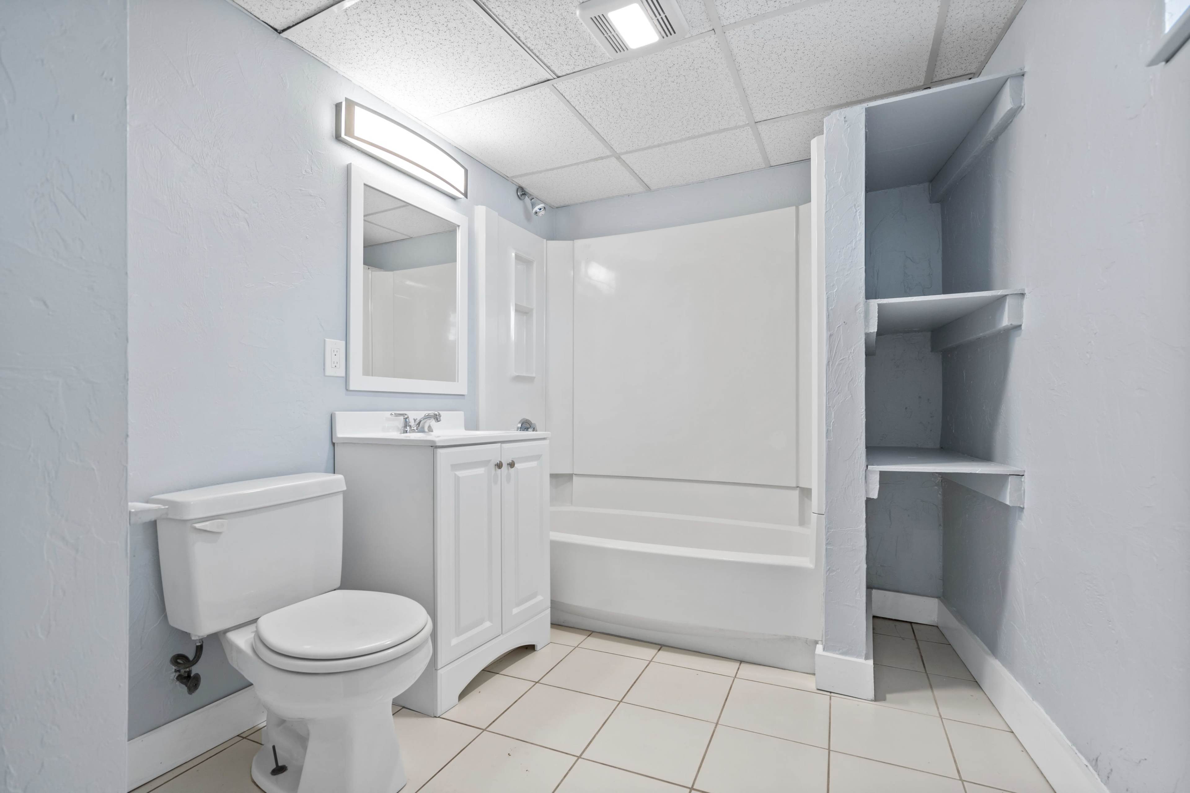 A bathroom features a toilet, a sink with a mirror, a bathtub, and wall-mounted shelves in a light blue environment.