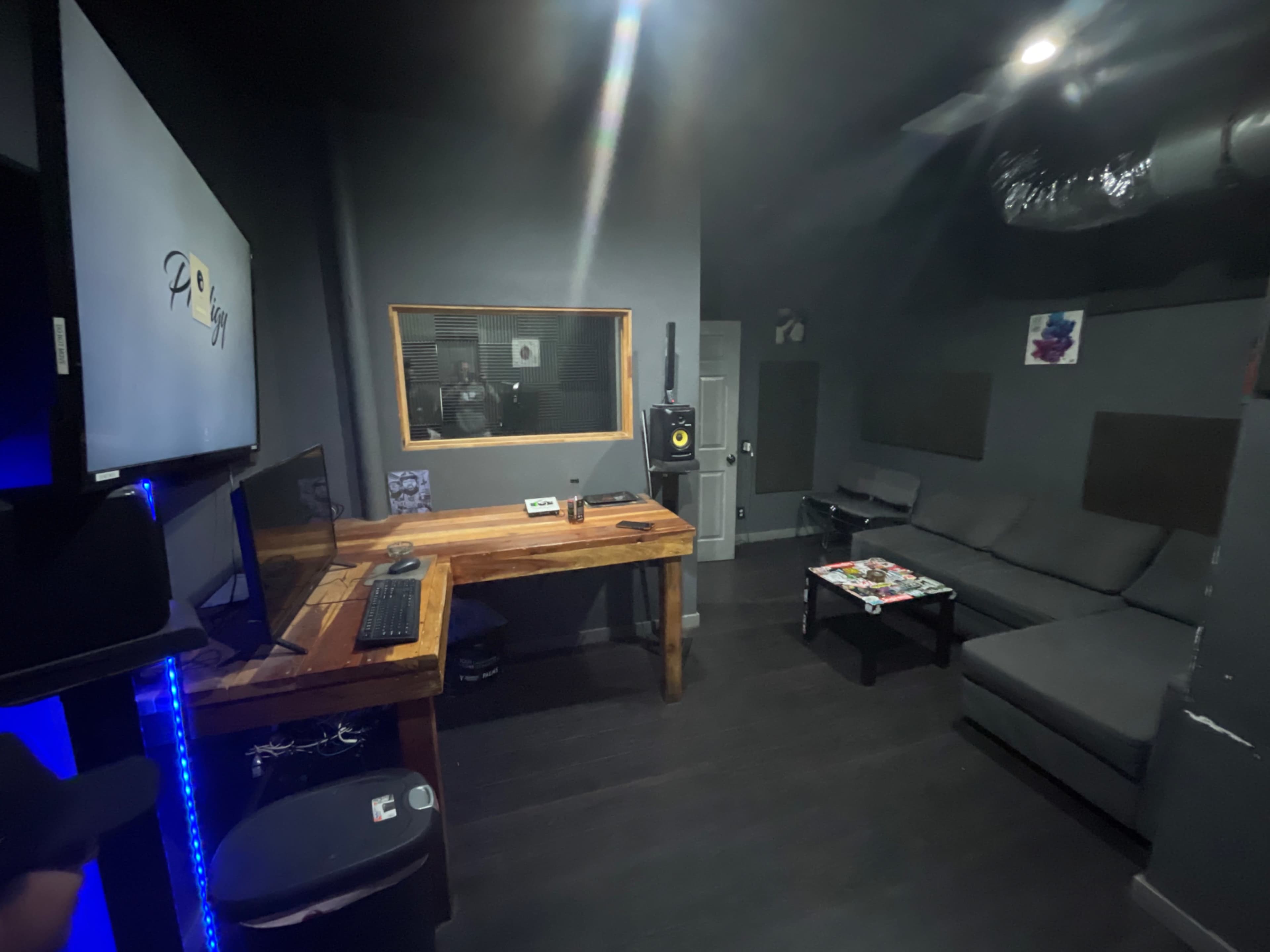 The image shows a music recording studio featuring a desk with a computer, a large soundproof window, a black sofa, and soundproofing panels on the walls.