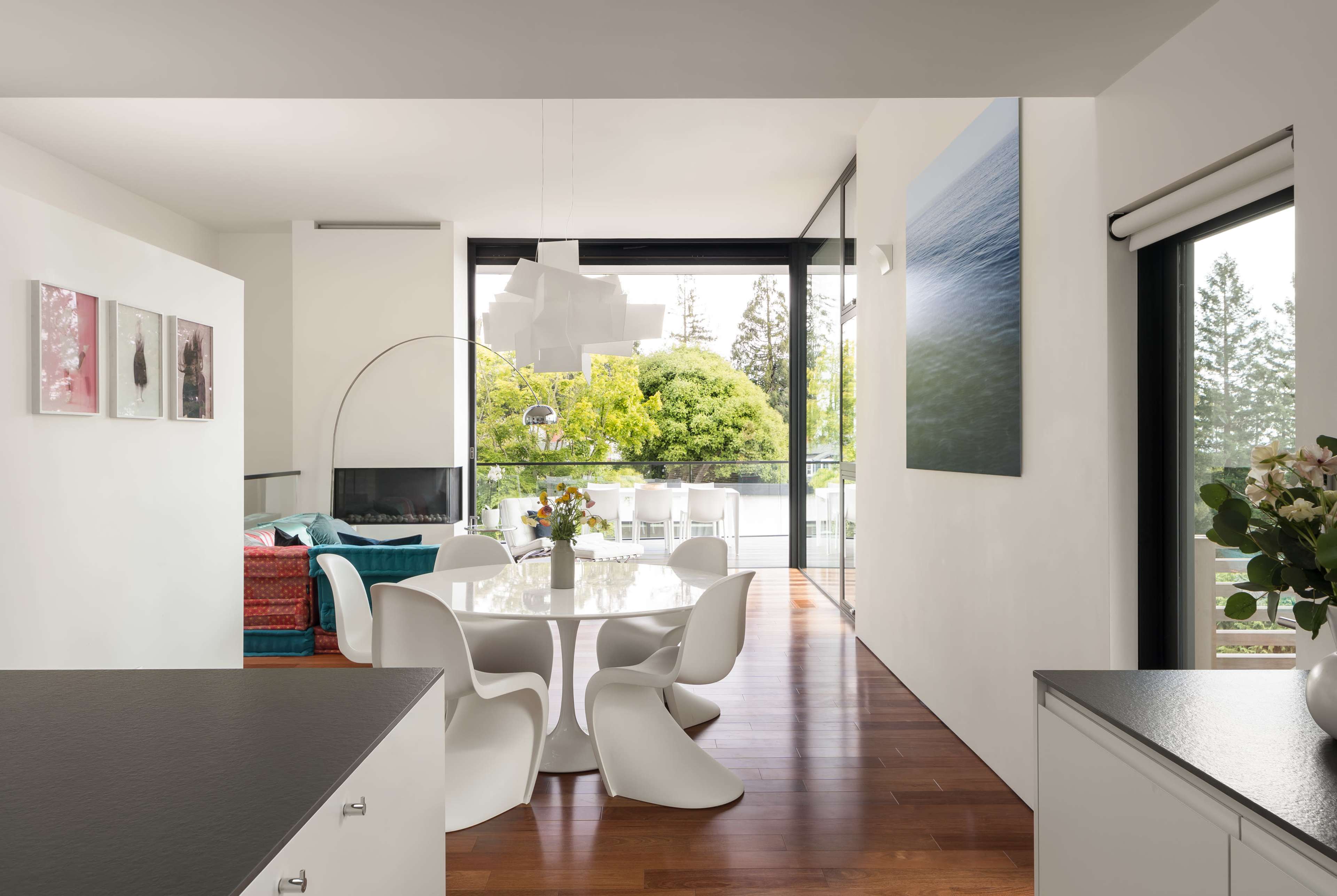 A bright, modern dining area features a circular white table surrounded by sleek chairs, with large windows providing a view of greenery outside.