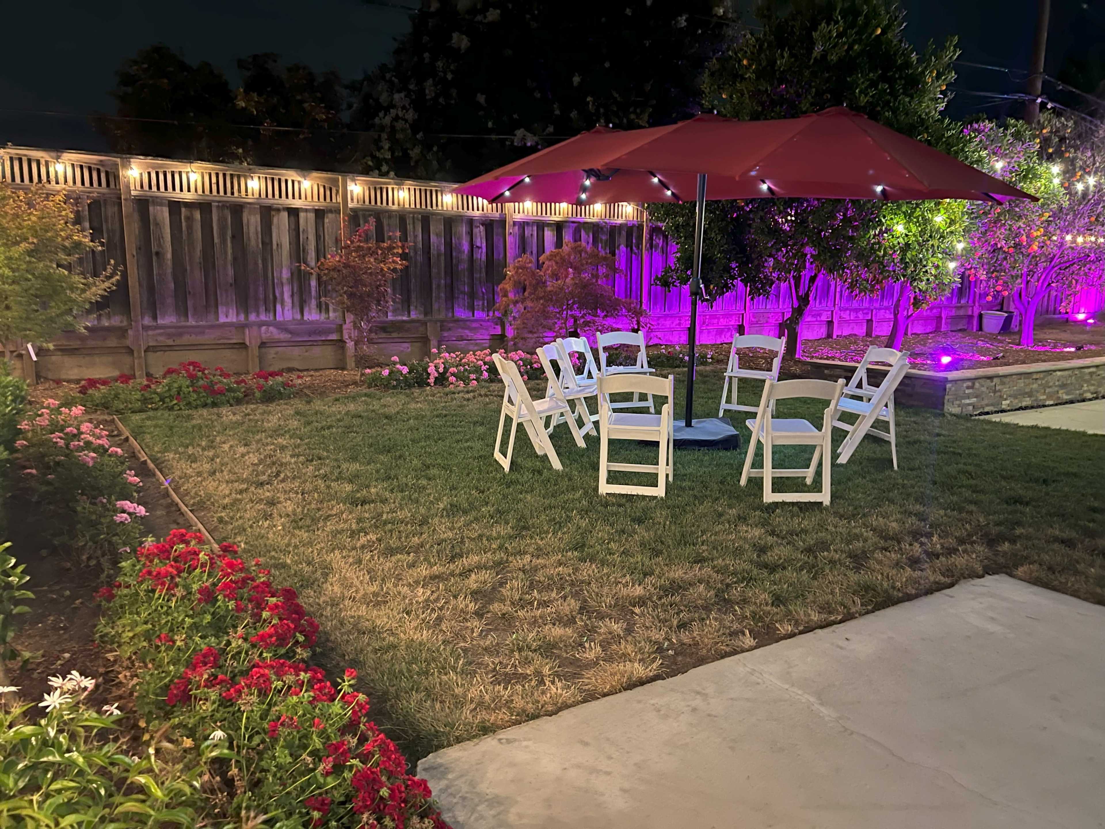 A circular arrangement of white chairs is set up under a red umbrella in a garden illuminated by purple lights.