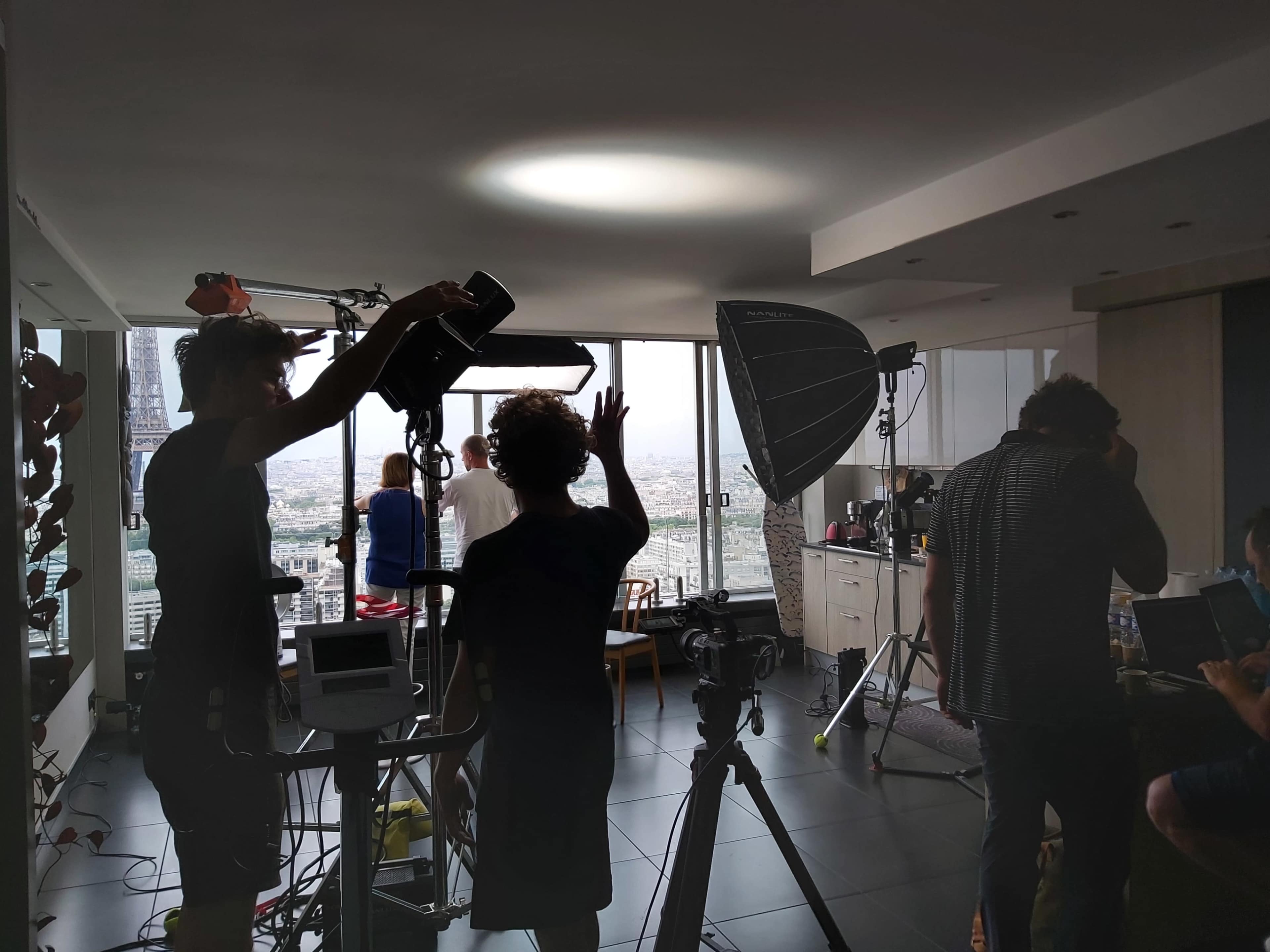 A film crew sets up equipment in a high-rise apartment with a view of the city.