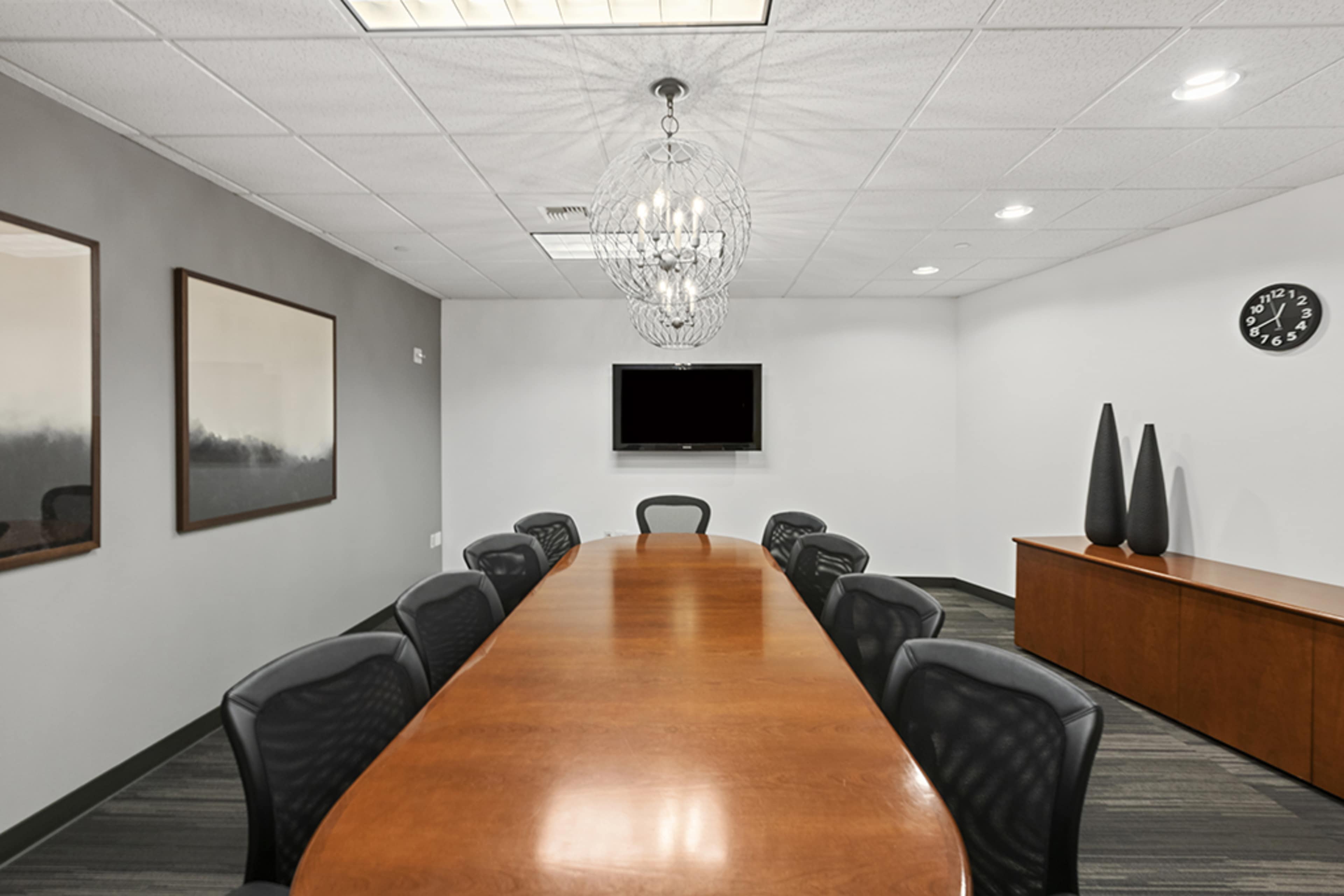 A long wooden conference table is arranged in a meeting room with black chairs, a large wall-mounted TV, and decorative elements such as lamps and framed artwork.