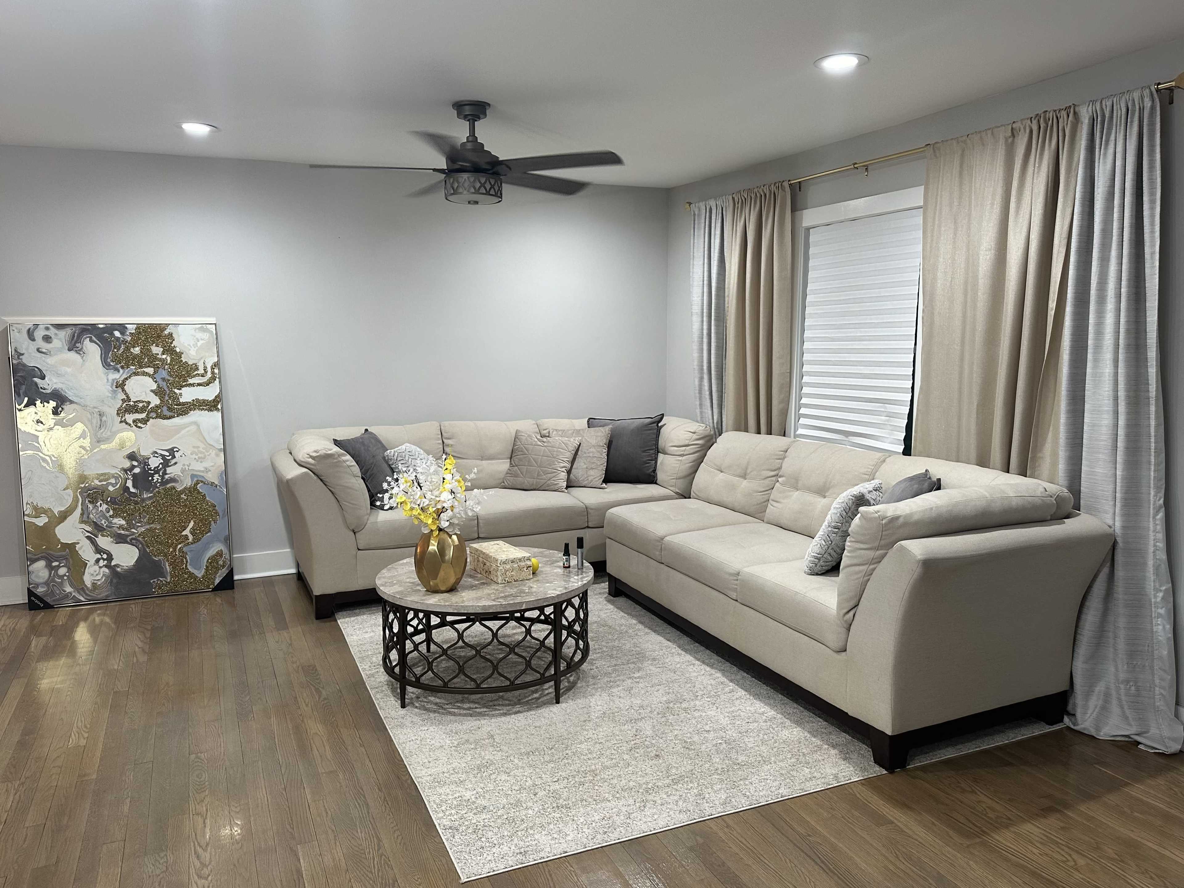 The living room features a sectional sofa, a round coffee table, and a wall art piece, all arranged on a hardwood floor with beige curtains framing a window.