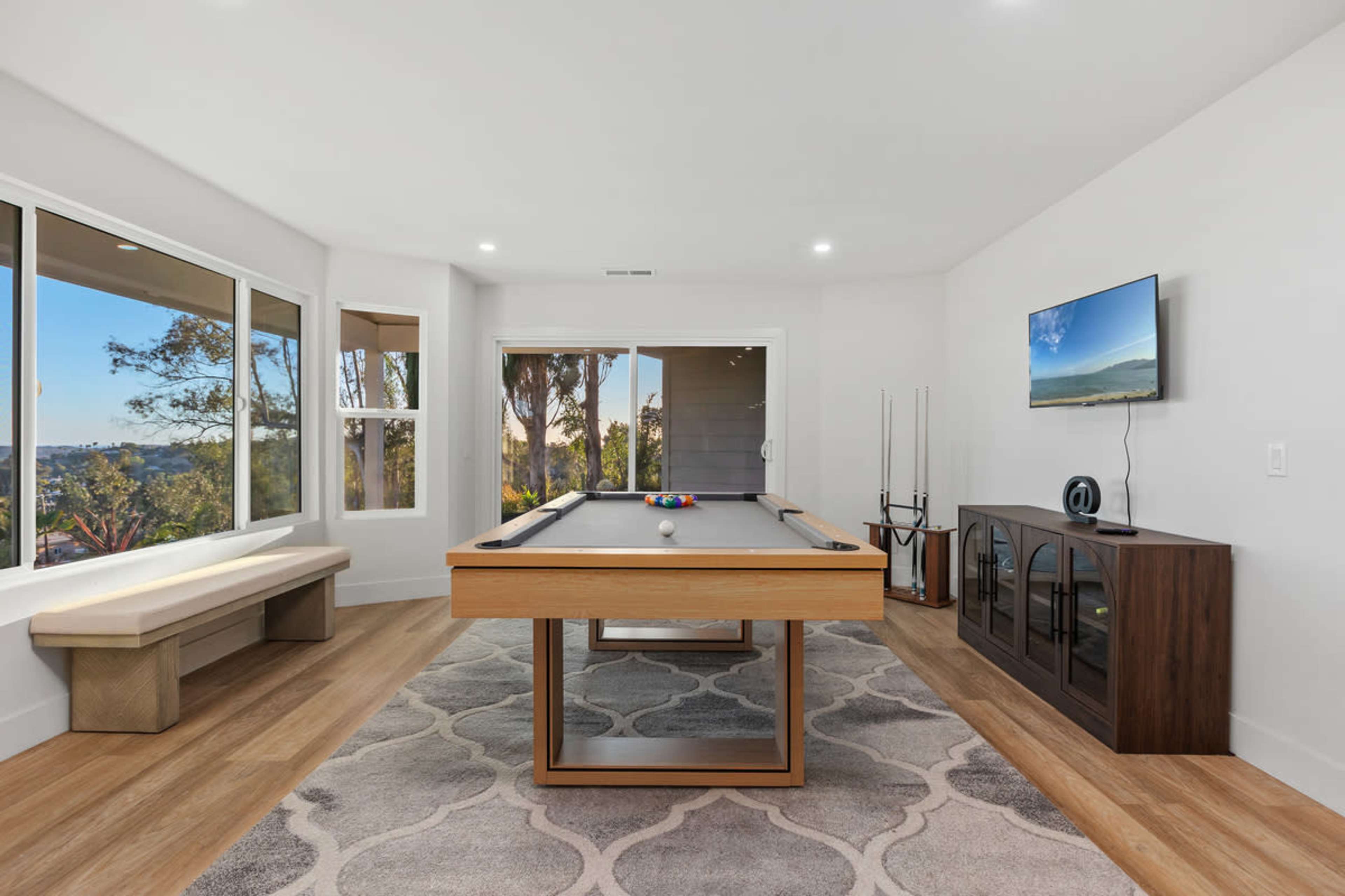 The image shows a modern game room featuring a pool table, large windows with outdoor views, and a TV mounted on the wall.