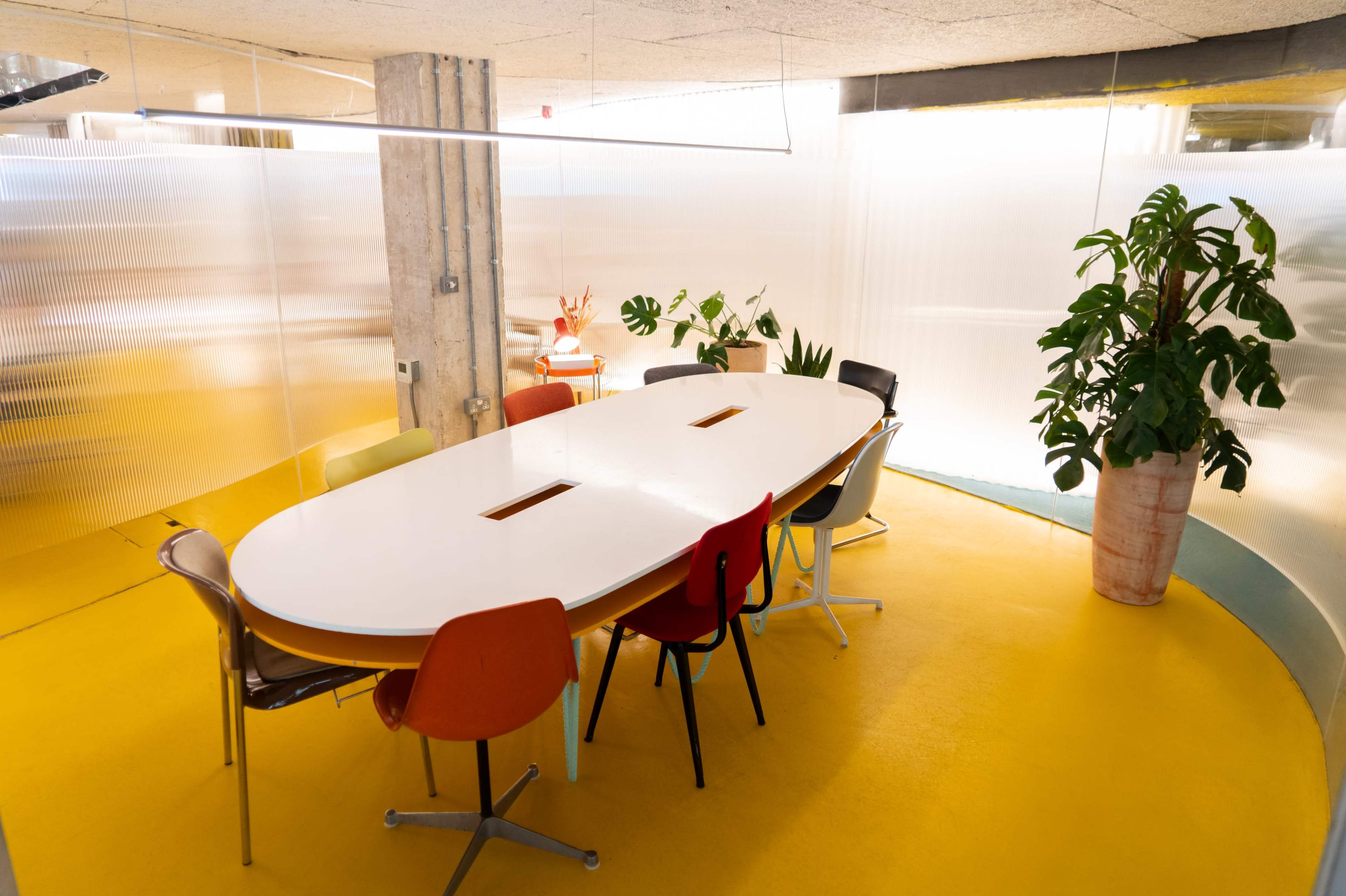 A large oval conference table is surrounded by colorful chairs in a modern office space with yellow flooring and green plants.