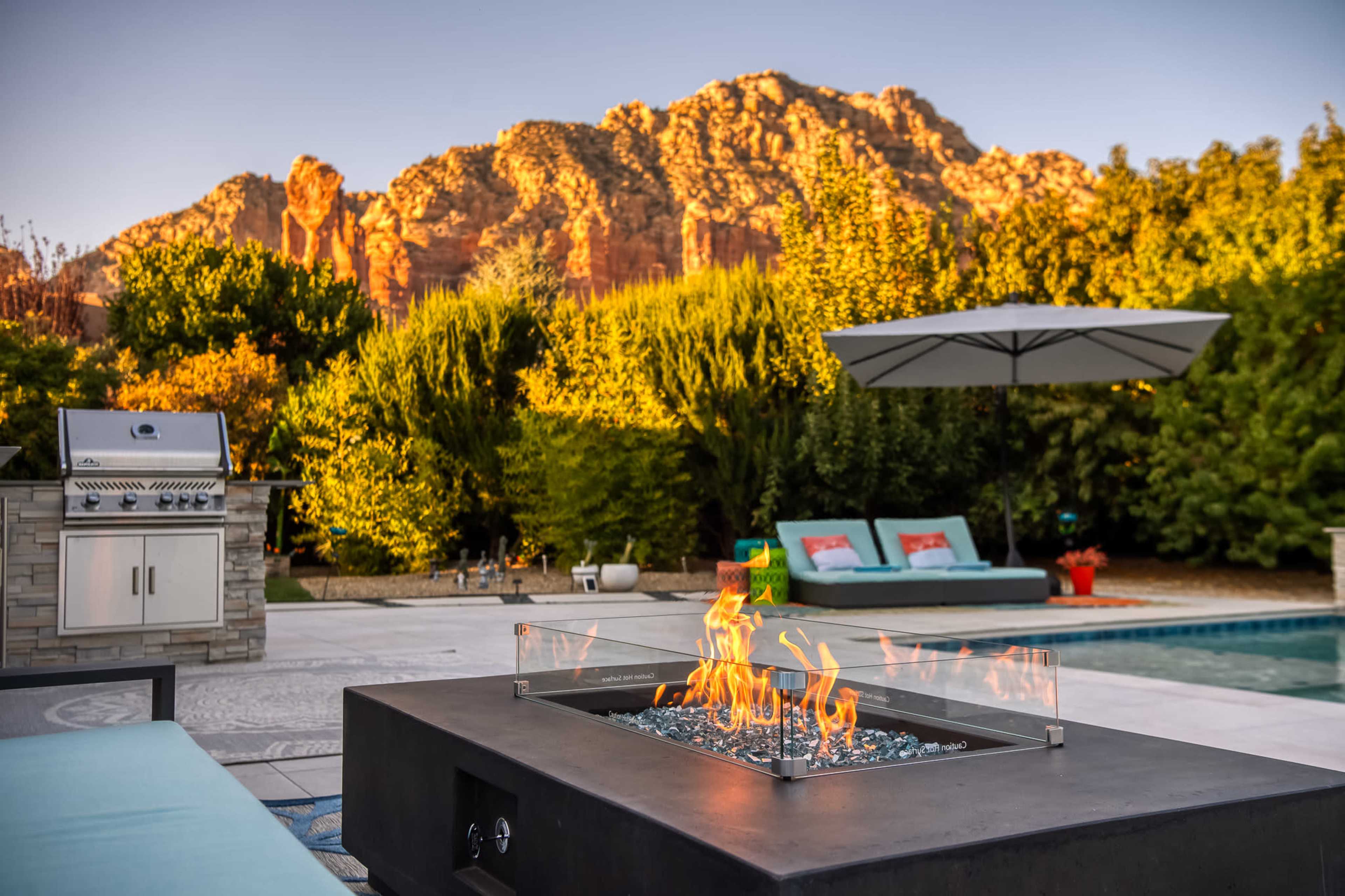 A fire pit with flames is situated in the foreground, surrounded by a stone patio, while a grill and lounge area are visible, with a backdrop of mountainous terrain and greenery.