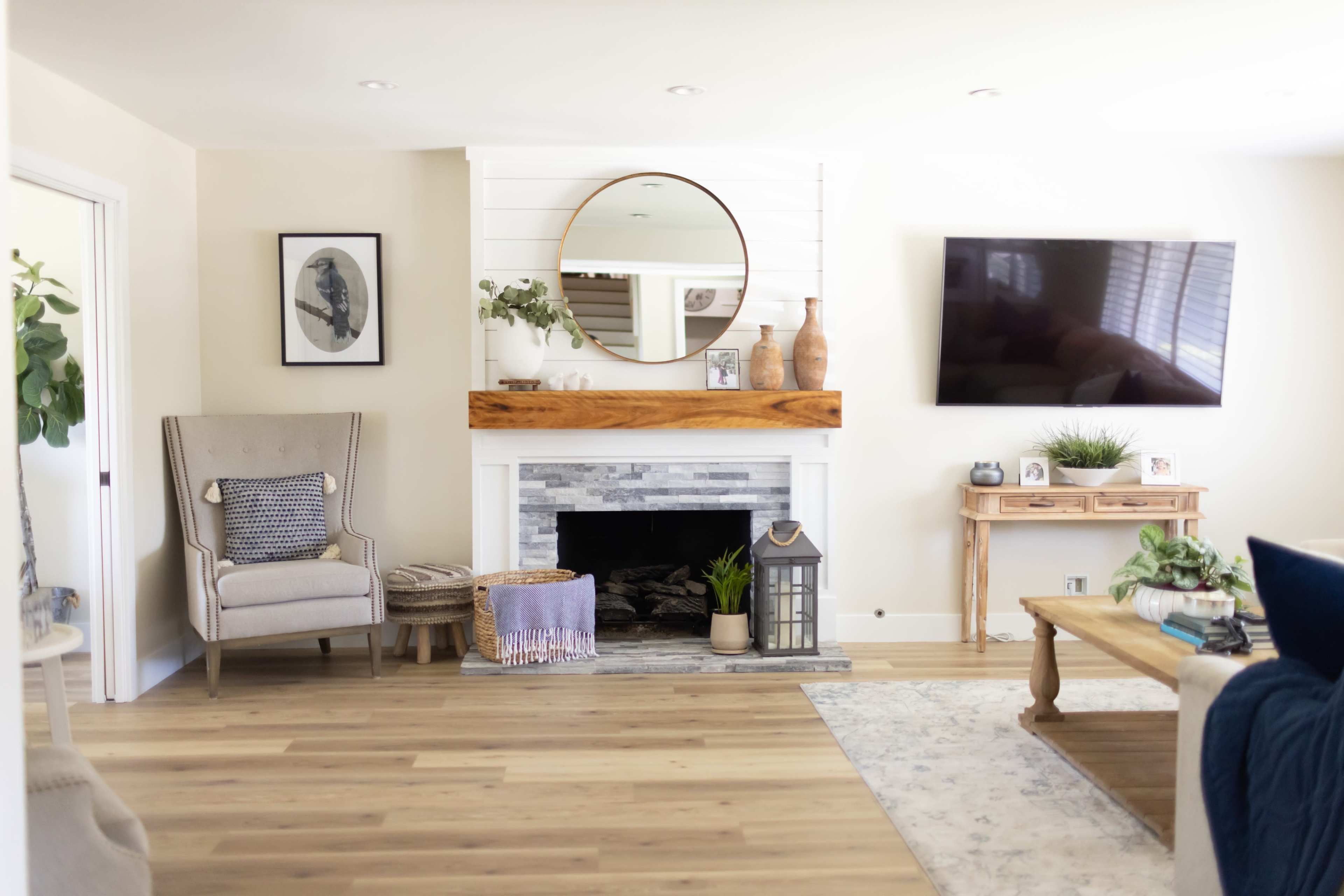 The living room features a fireplace with a wooden mantel, a round mirror above it, a television mounted on the wall, and a seating area with a chair and a decorative table.