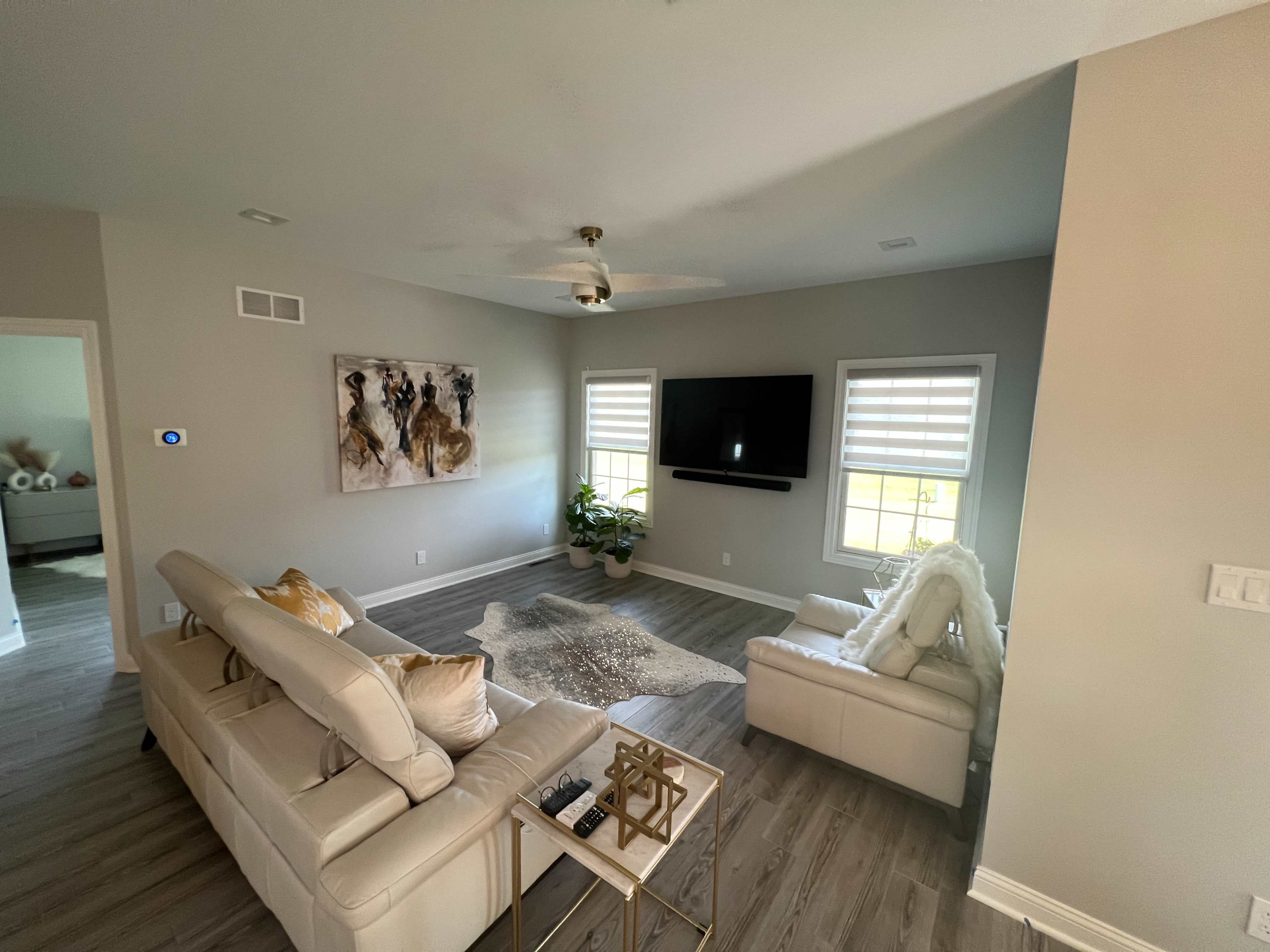 A modern living room features a beige sectional sofa, a large wall-mounted TV, and artwork alongside windows with white shutters.
