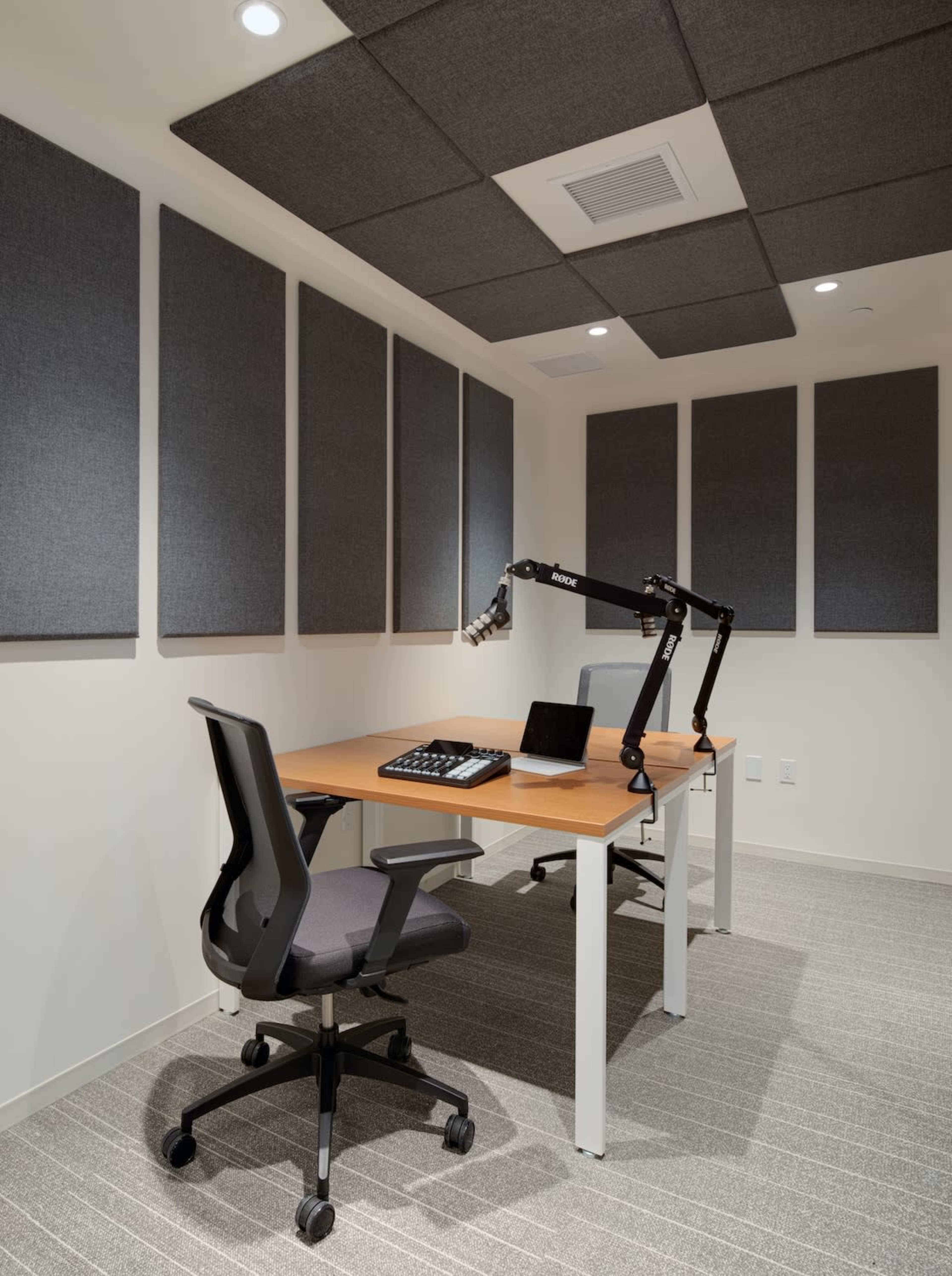 A minimalist office space featuring a large wooden desk, an adjustable microphone arm, a laptop, and acoustic panels on the walls.
