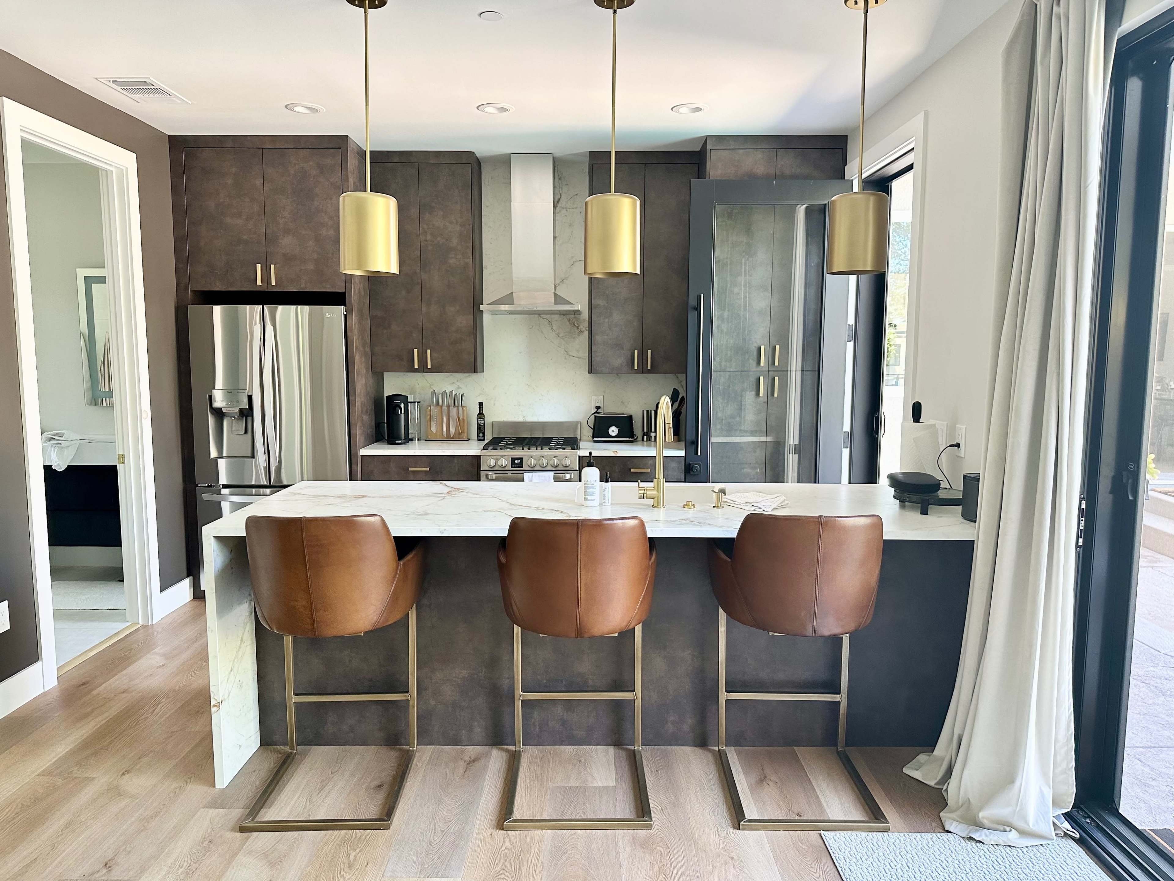 A modern kitchen featuring dark cabinetry, stainless steel appliances, and a marble-topped island with three brown leather bar stools.