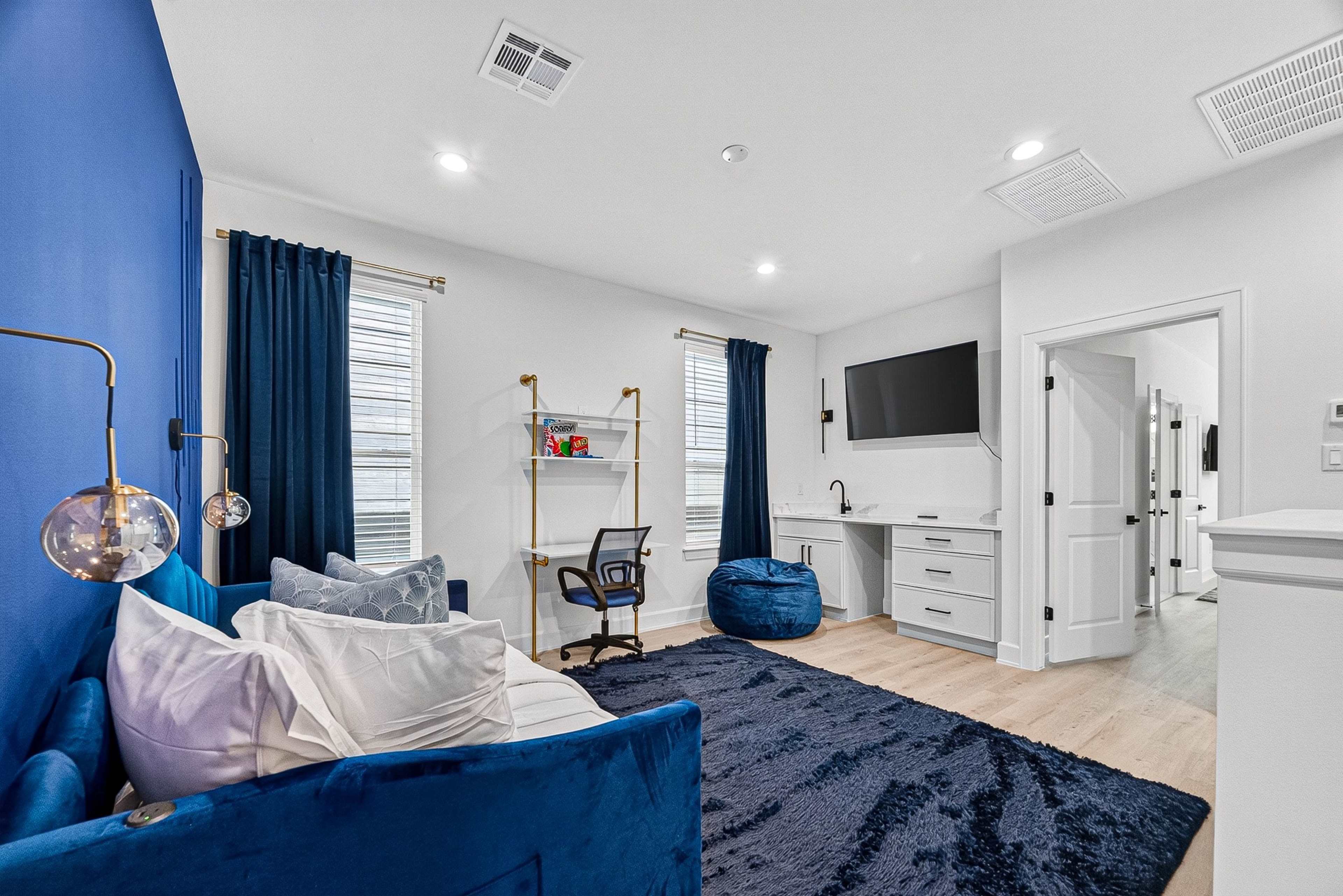 The image shows a modern room featuring a blue accent wall, a cozy sofa, a work area with a chair, and ample natural light from windows.