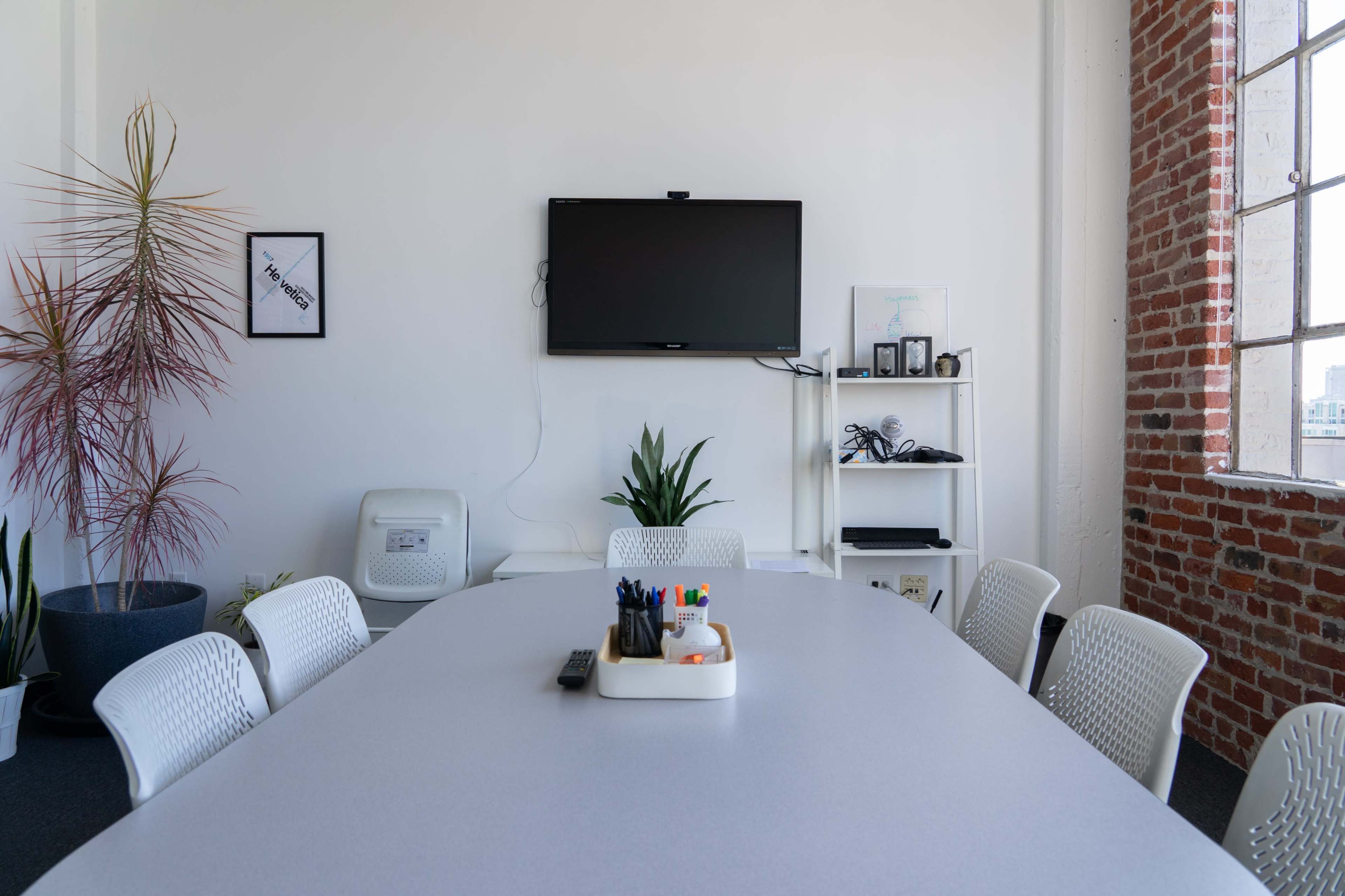 A conference room features a large table surrounded by white chairs, a television mounted on the wall, and a shelf with various items and plants.
